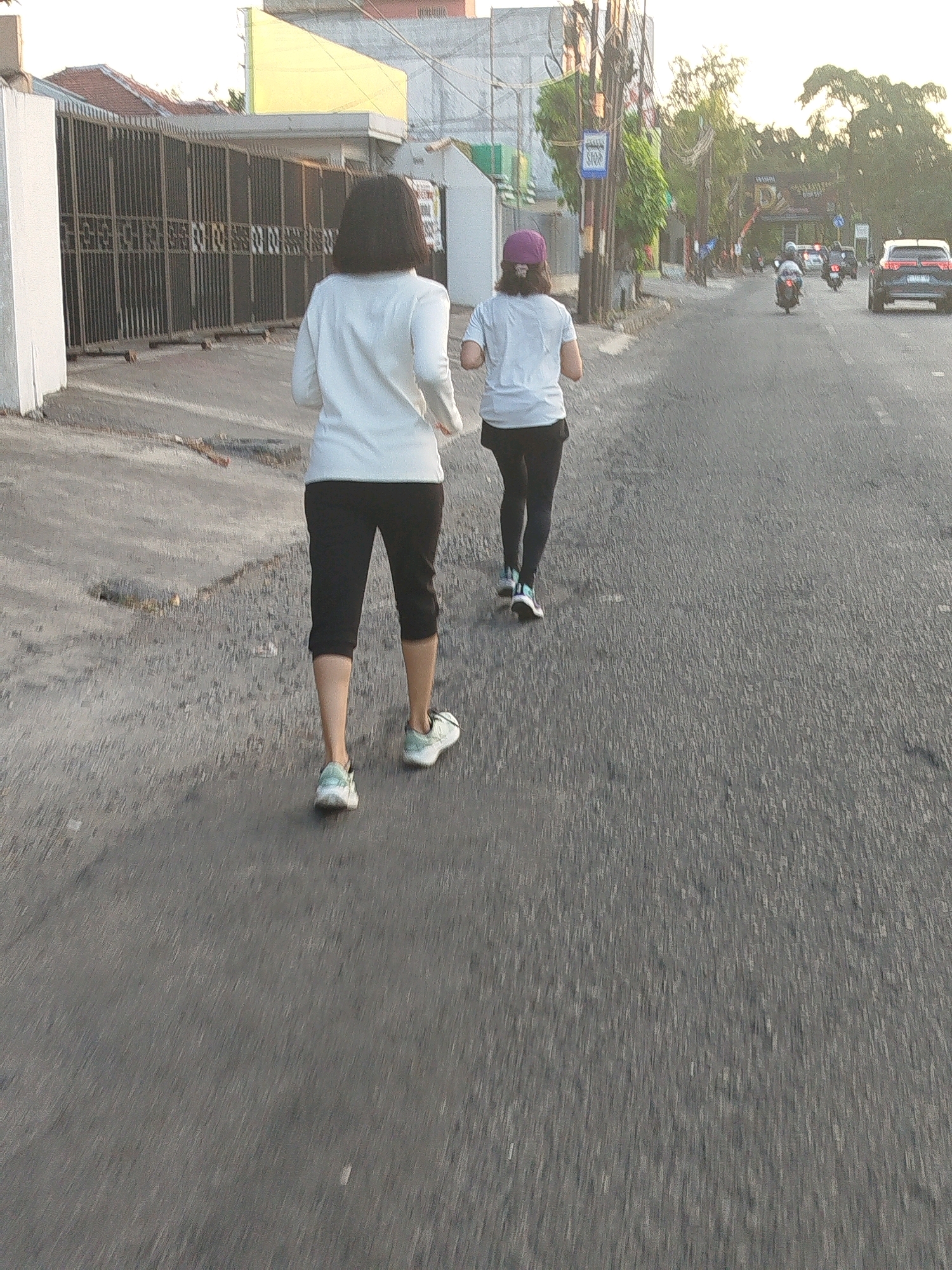 The image depicts two individuals running on a street in Surabaya, Indonesia. The person in the foreground, viewed from behind, is wearing a white long-sleeved shirt, black capri pants, and white athletic shoes with teal accents. The second person, also viewed from behind and slightly further ahead, is wearing a white t-shirt and dark pants, with a purple baseball cap. Both are running on an asphalt road. In the background, a dark-colored SUV is visible, along with several motorcycles and a bus stop sign. The scene appears to be in a residential or commercial area with buildings and a fence visible on the left side of the road. The overall lighting suggests it is either early morning or late afternoon.