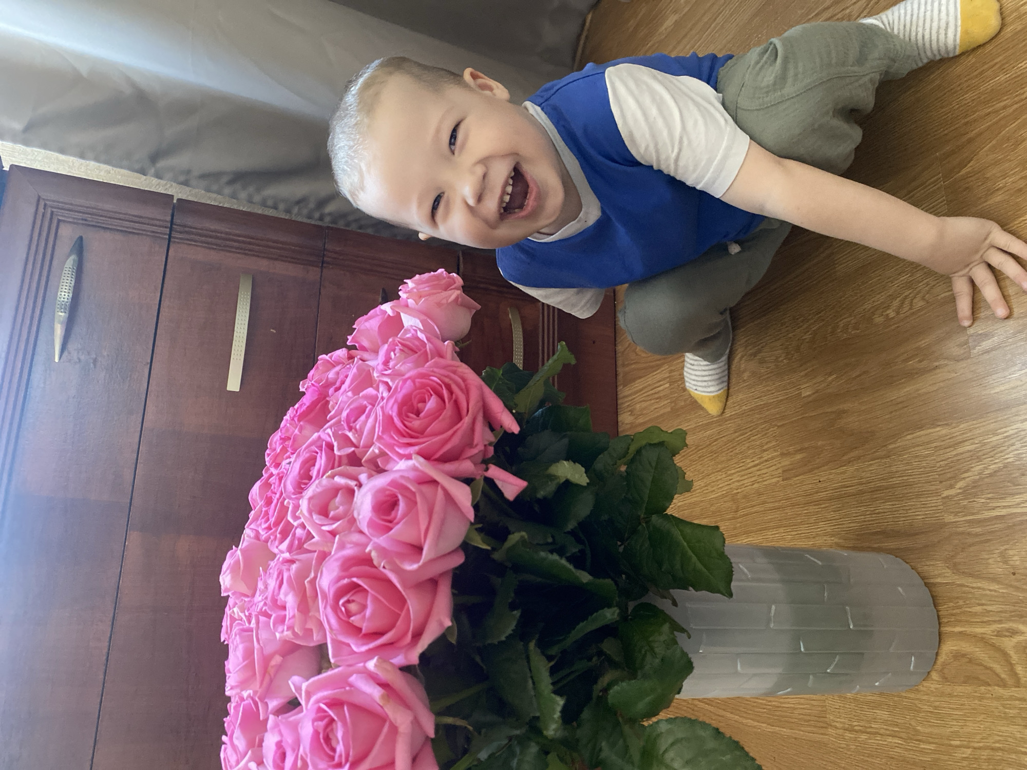 A bright-eyed toddler with a wide smile kneels on a wooden floor beside a large bouquet of pink roses. The child is dressed in a white t-shirt layered under a blue v-neck top, paired with olive-green pants and striped socks with yellow toes. The roses are in a light-colored, textured vase. Behind the roses is a dark wooden cabinet with metallic handles. A grey curtain is visible in the upper left corner. The scene appears to be indoors, potentially in a home. There is no information present in the image to identify Sochi, Russia, as the location.