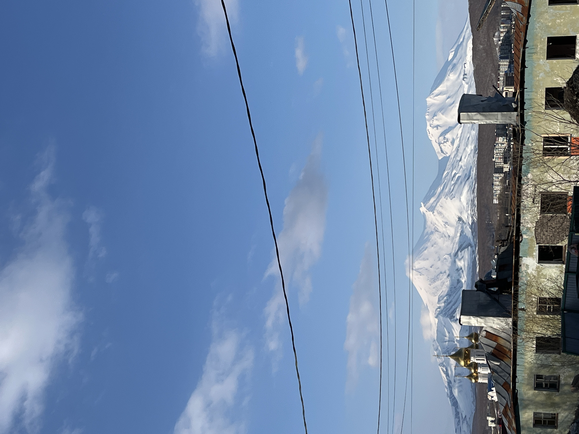 The image shows a view in Petropavlovsk-Kamchatsky, Russia, dominated by the snow-covered Vilyuchinsky volcano in the background. In the foreground are dilapidated buildings with open windows and rusting roofs. Power lines stretch across the sky. A golden-domed church is nestled among the buildings. The sky is blue with scattered clouds.
