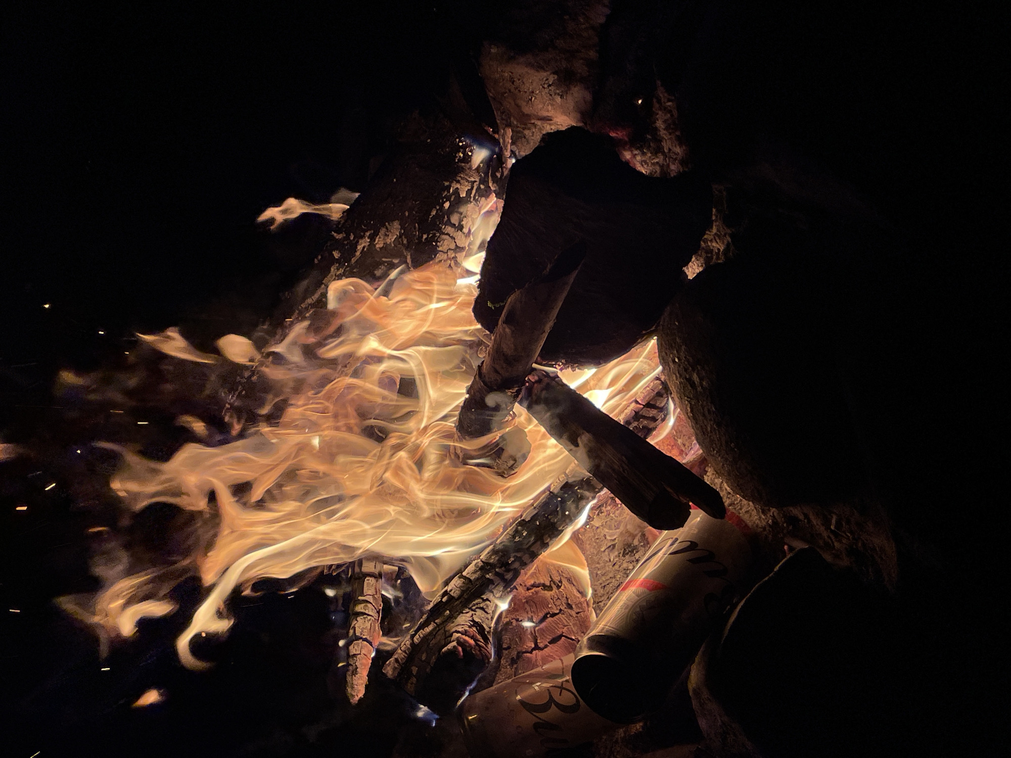 A close-up, low-angle shot captures a crackling campfire at night, set against a black background. The flames, rendered in warm hues of orange and yellow, dance and writhe, illuminating the surrounding logs and embers. Several logs are visible, some appearing charred and others glowing with heat. To the left, two aluminum cans, one partially obscured, are nestled among the rocks and wood. The cans appear to be of a beverage brand. The scene suggests a rustic outdoor setting, possibly a campsite. No people are visible. The image is devoid of any identifying markers of Berozovo, Russia.