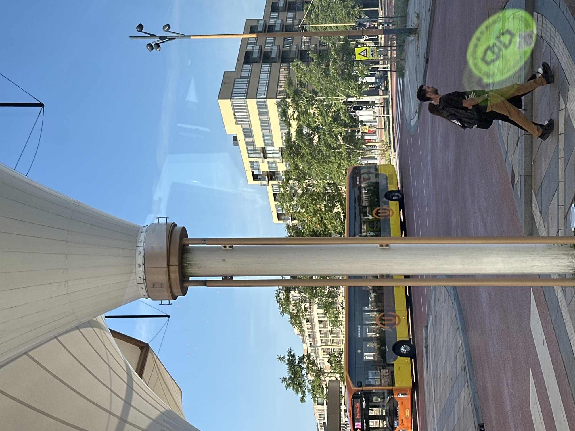 The image captures a street scene in Utrecht, Netherlands, dominated by a modern building with a yellow facade, lush green trees, and a yellow and grey bus. In the foreground, a large white and beige canopy structure with metal supports is partially visible, casting shadows. Two individuals, a man and a woman, are walking across a crosswalk. The man is wearing a black t-shirt and khaki pants, while the woman is wearing a black top and pants, carrying a backpack. A bright green, projected graphic of a stylized bicycle is visible on the pavement near them. The street is paved with red asphalt, and there are traffic signals and streetlights in the background. The sky is clear and blue.