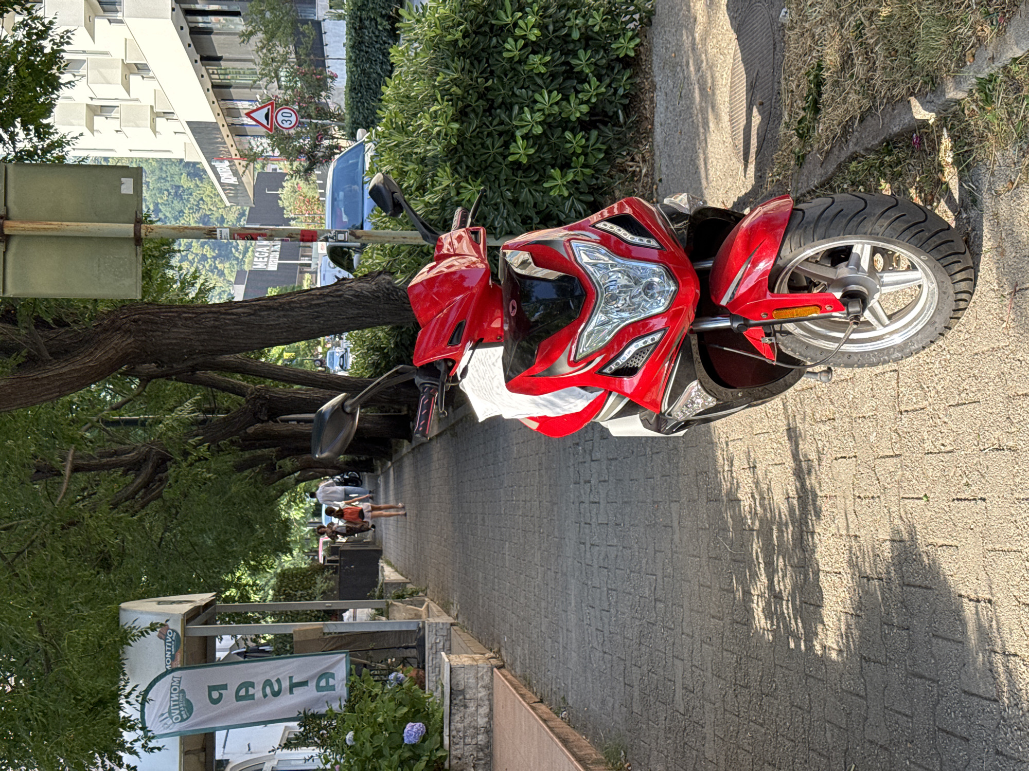 The image depicts a street scene in Budva, Montenegro, with a red scooter prominently parked on the right side of a paved pedestrian walkway. The walkway is bordered by trees casting dappled shadows on the ground. On the left, a restaurant with a sign reading "PASTA" is visible, along with lush green plants. Further down the walkway, several people are walking, including a woman with a red backpack and a man in a white shirt. In the background, buildings and vehicles can be seen, along with a traffic sign indicating a speed limit of 30. The overall atmosphere suggests a sunny day in a European coastal town.