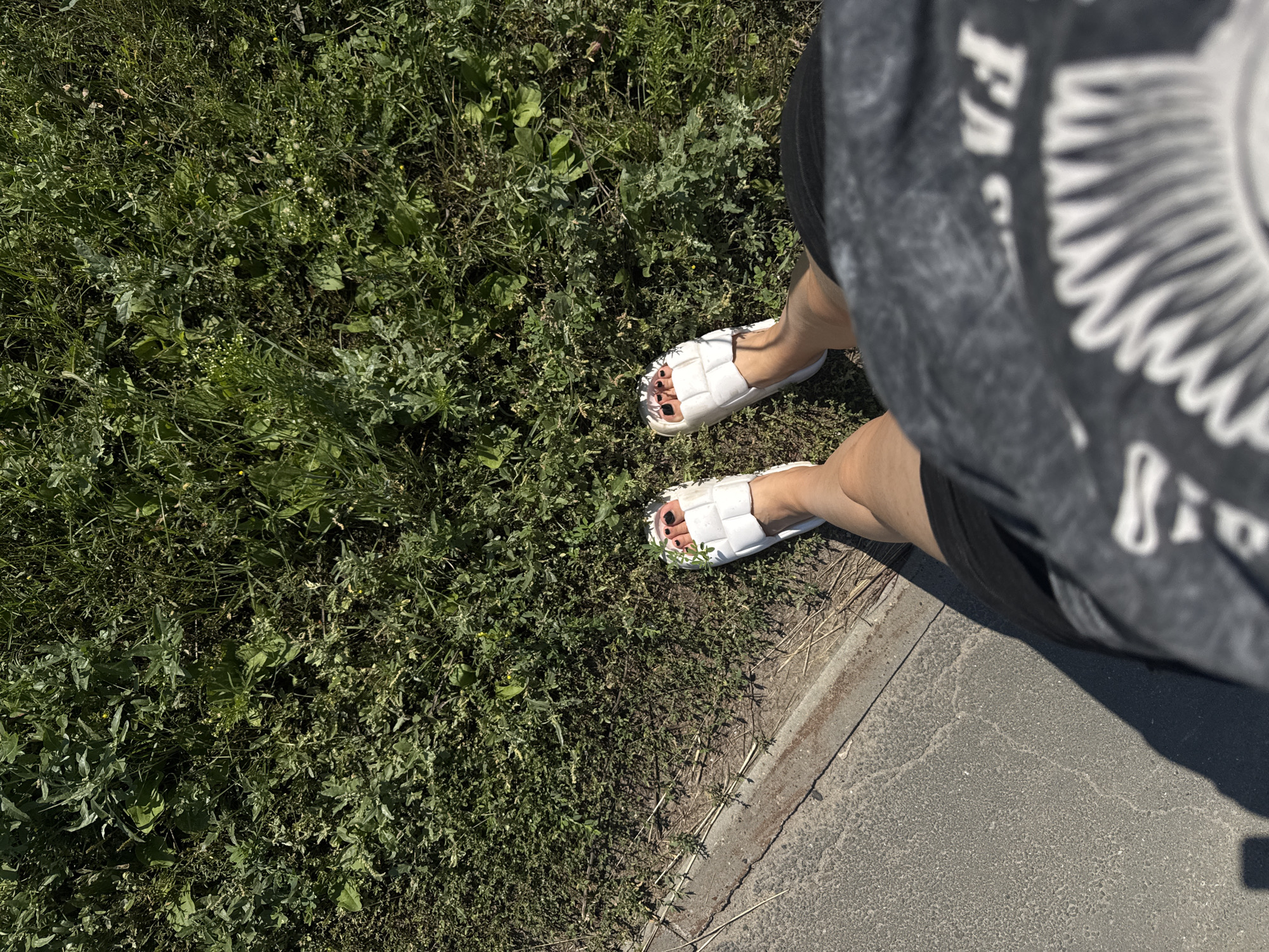 The image shows a person's legs and feet, wearing white slides and dark shorts, standing on a paved path next to a grassy area. The grass is dense and appears to be a mix of various plants and weeds. The lighting suggests it is daytime and sunny. The image is taken from a high-angle perspective, looking down. The location is identified as the city of Tolyatti, Russia.