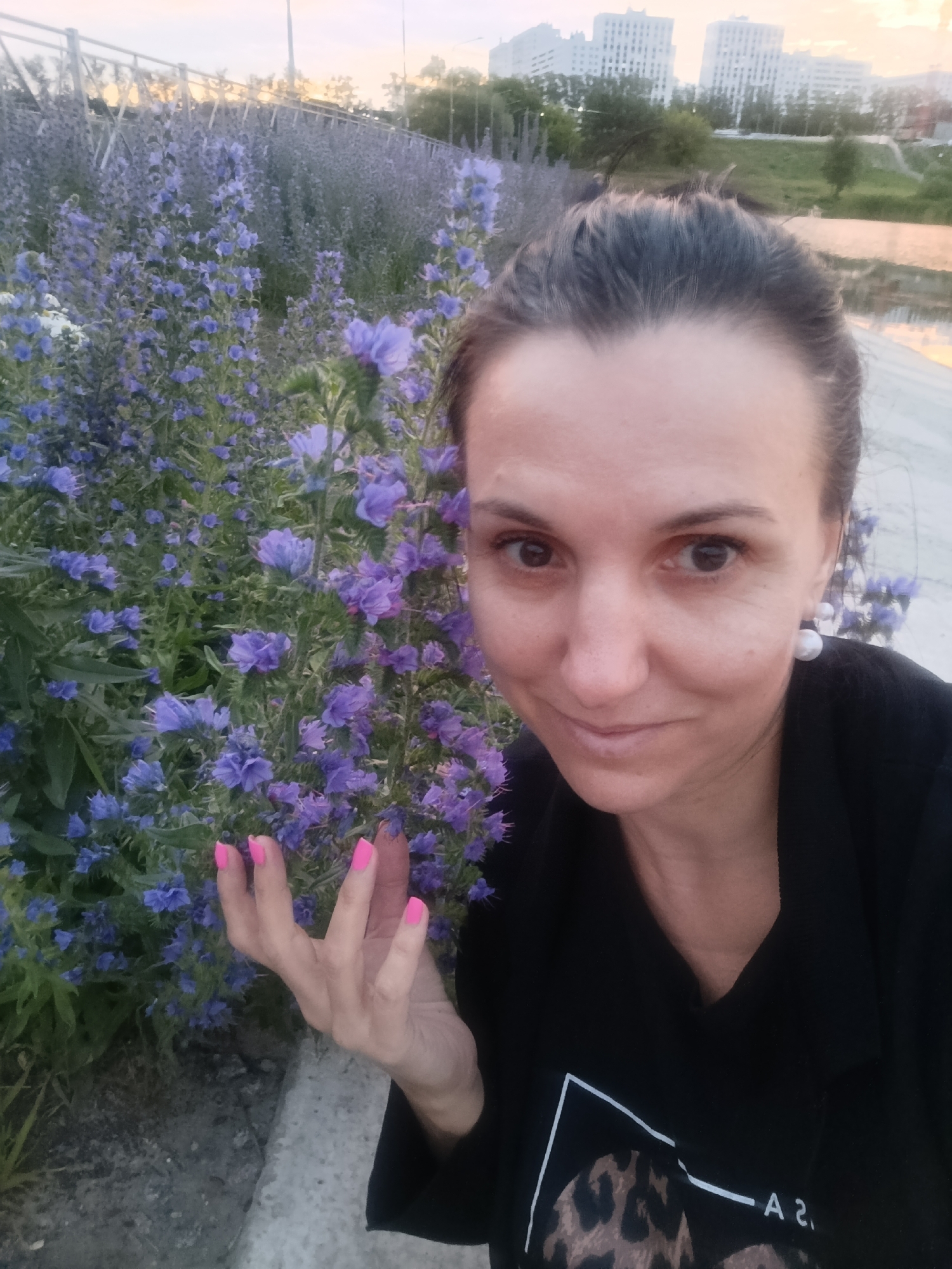 A woman with dark hair pulled back, wearing a black top with a V-neck and a leopard print design on the front, poses for a selfie outdoors. She has on large pearl earrings and bright pink nail polish. The background is filled with lush, tall blue wildflowers and a stone curb. In the distance, modern white buildings rise against a pale sky, suggesting an urban environment. The overall lighting indicates it is either dawn or dusk. The location appears to be a park or a green space within the city of Penza, Russia, given the presence of the wildflowers and the urban background.