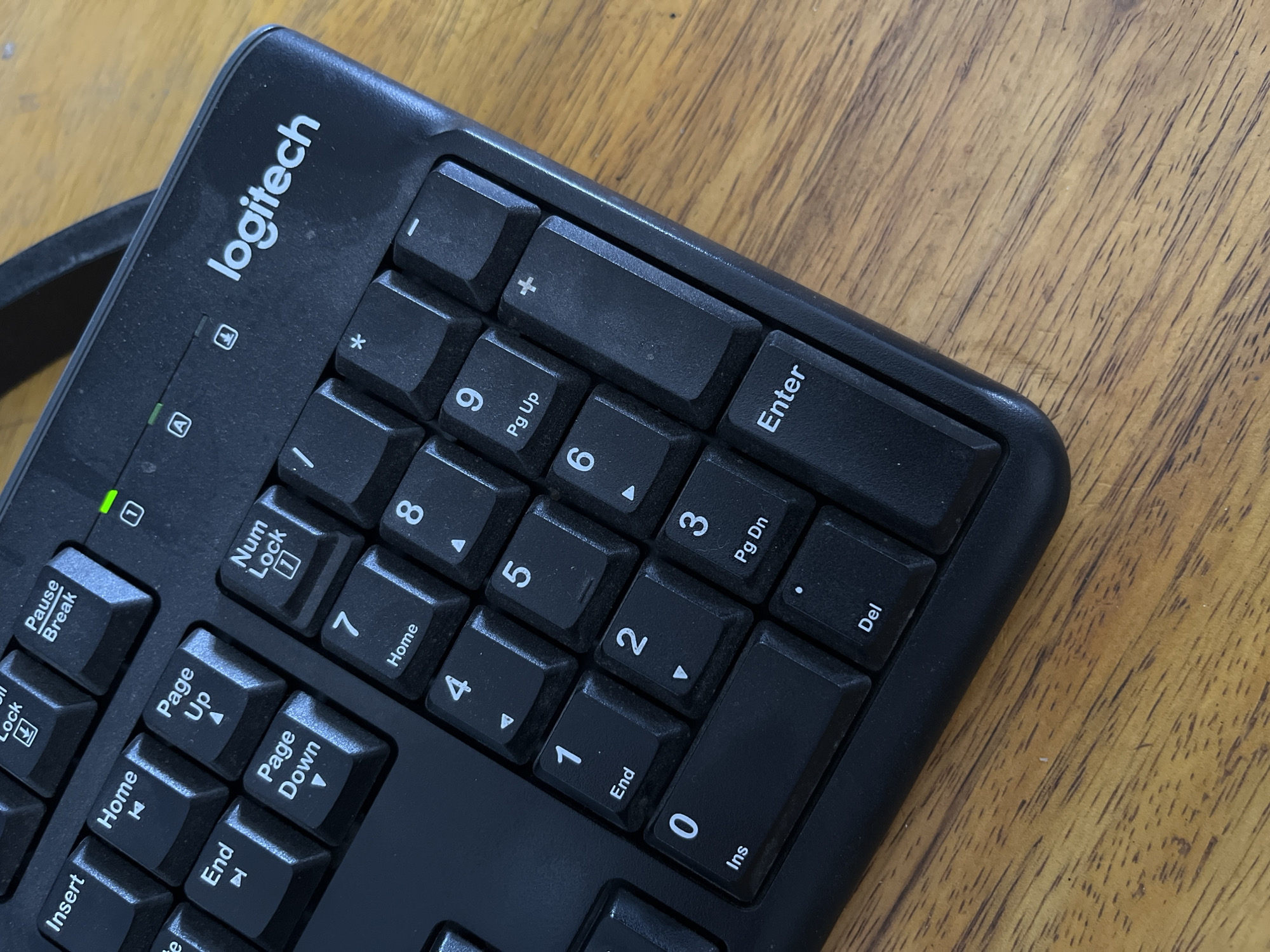The image shows a close-up, overhead view of a black Logitech keyboard resting on a wooden surface, likely a table. The keyboard is positioned at an angle, with the right side and top edge of the number pad prominently displayed. Several keys are visible, including "Insert", "Home", "Page Up", "Page Down", "End", "Num Lock", number keys from 0 to 9, ".", "Enter", and "+". The "Logitech" logo is visible in white lettering on the top right of the keyboard. A green LED indicator light is also visible above the "Num Lock" key. The wooden surface appears to be a light to medium brown with visible wood grain. There are no people, animals, or any other identifiable objects present in the scene. The environment is static and appears to be indoors, given the keyboard and wooden table. The image is well-lit, with a natural light source casting subtle shadows. The location of Kedawung, Indonesia, is not discernible from the visual information presented in the image.
