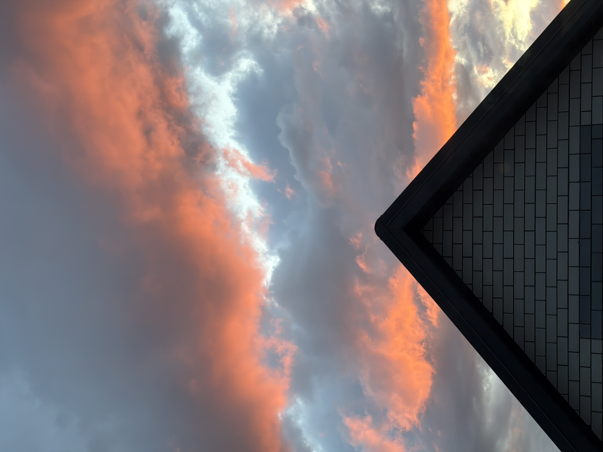 The image captures a view of a sunset sky with orange and grey clouds, juxtaposed with the corner of a building featuring brickwork and dark trim. The building's corner is positioned on the right side of the frame, while the sky occupies the majority of the left. The brickwork appears to be grey, and the trim is a dark color, possibly black. The location is Rossiyskiy, Russia.