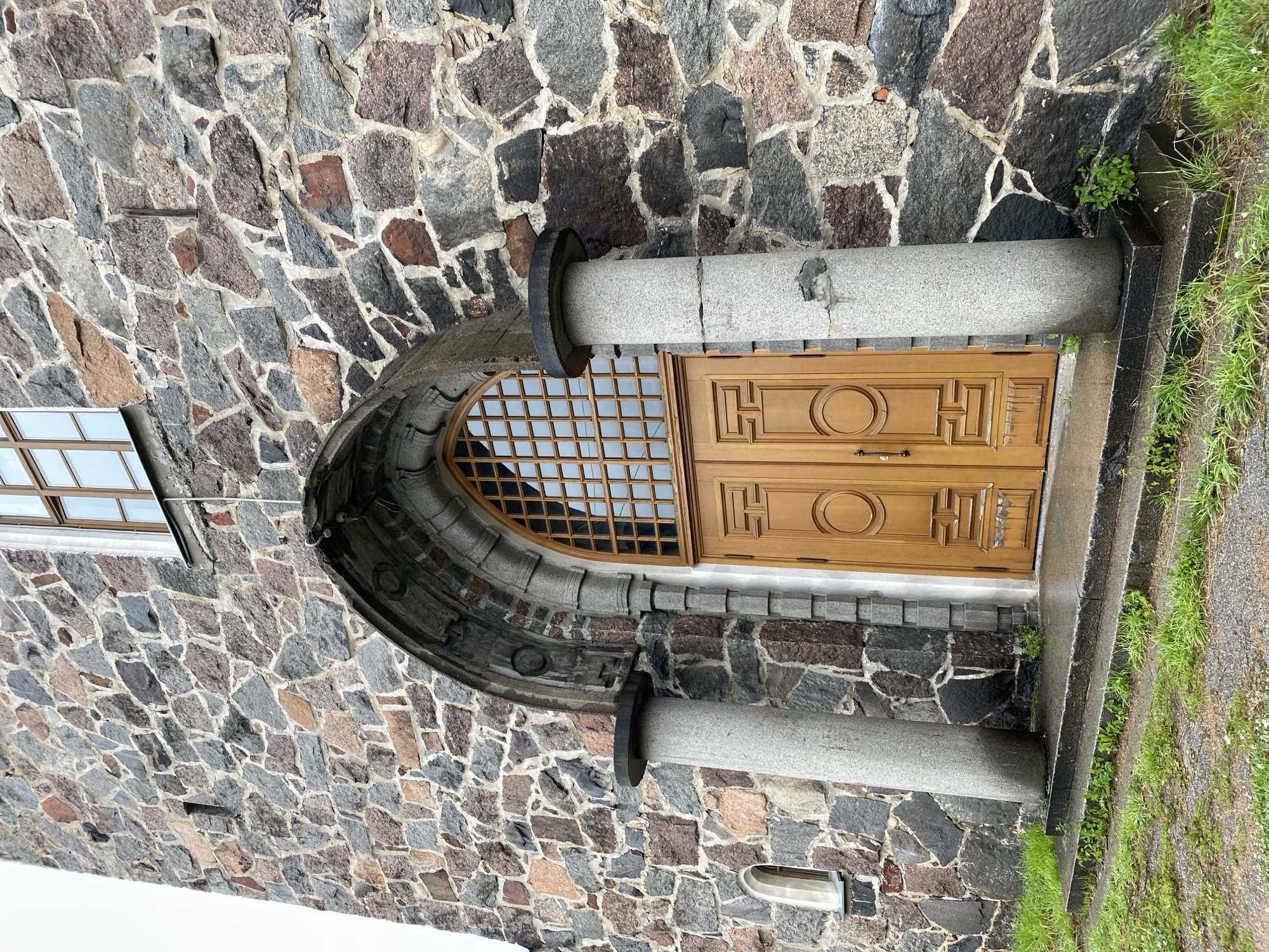 The image shows the stone facade of a building, likely a church, in Priozersk, Russia. A large, arched entryway with a double wooden door dominates the center of the frame. The door is made of light-colored wood and features decorative circular and geometric panels. Above the door is a large, arched window with a grid of wooden panes. Two thick, cylindrical stone pillars stand on either side of the entrance, supporting a decorative stone archway. The walls of the building are constructed from rough-hewn stones of various sizes and colors, including gray, pink, and dark brown. A small, narrow window is visible on the left side of the facade. The ground in front of the entrance is covered with a few stone steps and patches of green grass and vegetation. The overall impression is of an old, sturdy stone structure.