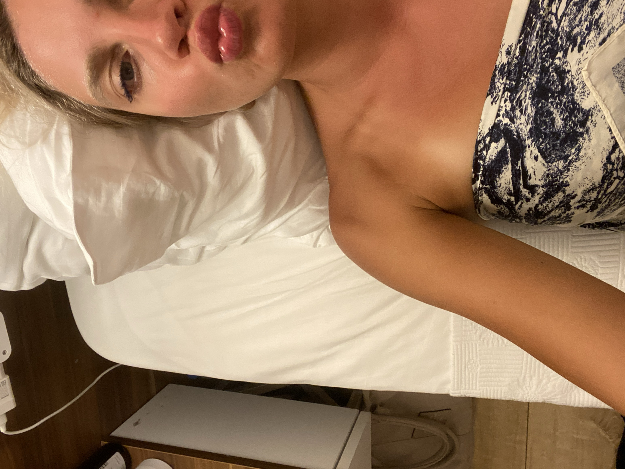 An overhead medium shot captures a young Caucasian woman lying in bed with her face in the upper right corner of the frame. Her eyes are closed, and her lips are pursed in a kiss. Her blonde hair is visible at the top of her head. The woman is wearing a strapless top with a blue and white abstract pattern. Her tanned skin is visible on her shoulder and arm. The bed is covered with white linens, and a white pillow is visible behind her head. To the left of the bed, a wooden nightstand is visible with a white object plugged into an electrical outlet. The background is dark and out of focus. This image was taken in the city of Aksu, Turkey.