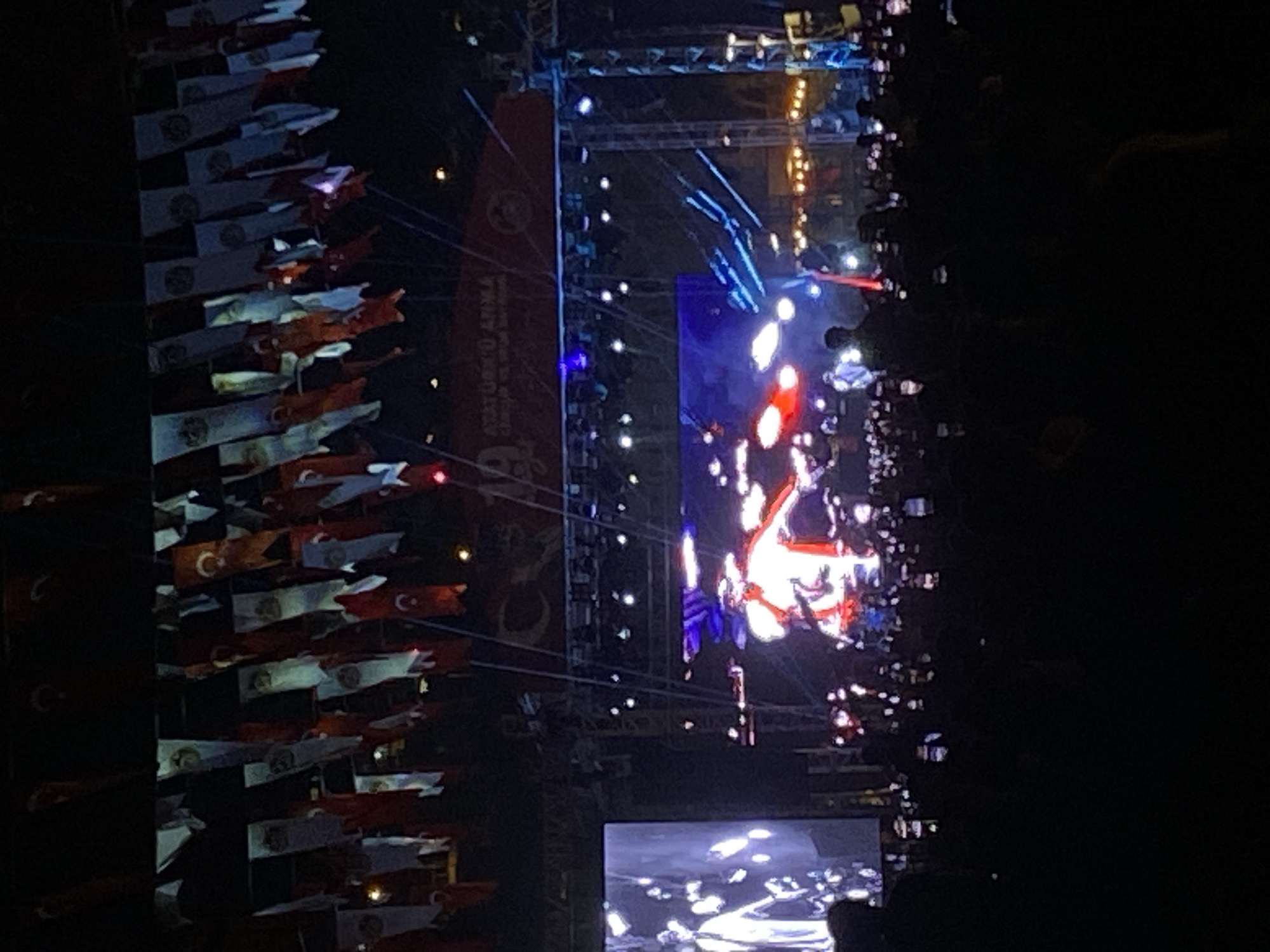 The image depicts a nighttime view of an event, possibly a performance or ceremony, taking place at the Alanya Ataturk Square in Alanya, Turkey. Numerous Turkish flags are visible, along with what appear to be spectators. A large screen displays imagery of bright, indistinct content, possibly related to the event. Stage lighting and rigging are also visible, suggesting a production or performance setting. The overall scene has a dynamic feel with bright light sources set against a dark background.
