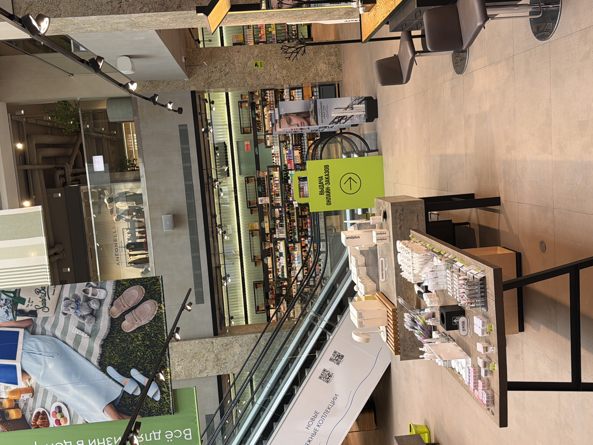 The image depicts the interior of a large, multi-level retail establishment, likely a department store or a shopping mall, with elements that suggest it is located in Moscow, Russia.
In the foreground, a wooden display table is laden with various cosmetic products, including numerous small bottles, jars, and tubes, predominantly in white and pastel packaging. A black cube-shaped container and a sign reading "УХОД" (which translates to "CARE" in Russian) are also visible on this table. To the right, a counter with a wooden top is equipped with two brown bar stools and storage drawers.
Dominating the midground is a brightly colored, neon green sign with a white arrow pointing to the right, accompanied by Russian text that reads "ВЫДАЧА ОНЛАЙН-ЗАКАЗОВ" ("ONLINE ORDER PICKUP"). Adjacent to this sign are several tall, narrow display stands featuring images and text related to beauty products, including a prominent advertisement for L'Oréal. Further back, an escalator slopes upwards, and a large banner featuring a person enjoying a picnic, along with text in Russian ("Всё для жизни в центр"), hangs from the upper level. The background reveals shelves stocked with numerous products, suggesting a large beauty or cosmetics section.
The architecture features exposed concrete pillars and a multi-story layout with balconies and railings. Track lighting illuminates the space. The floor is tiled in a neutral, large-format pattern.
The overall impression is a clean, modern retail environment focused on beauty and consumer goods. The presence of Russian language signage definitively places the location in Russia.