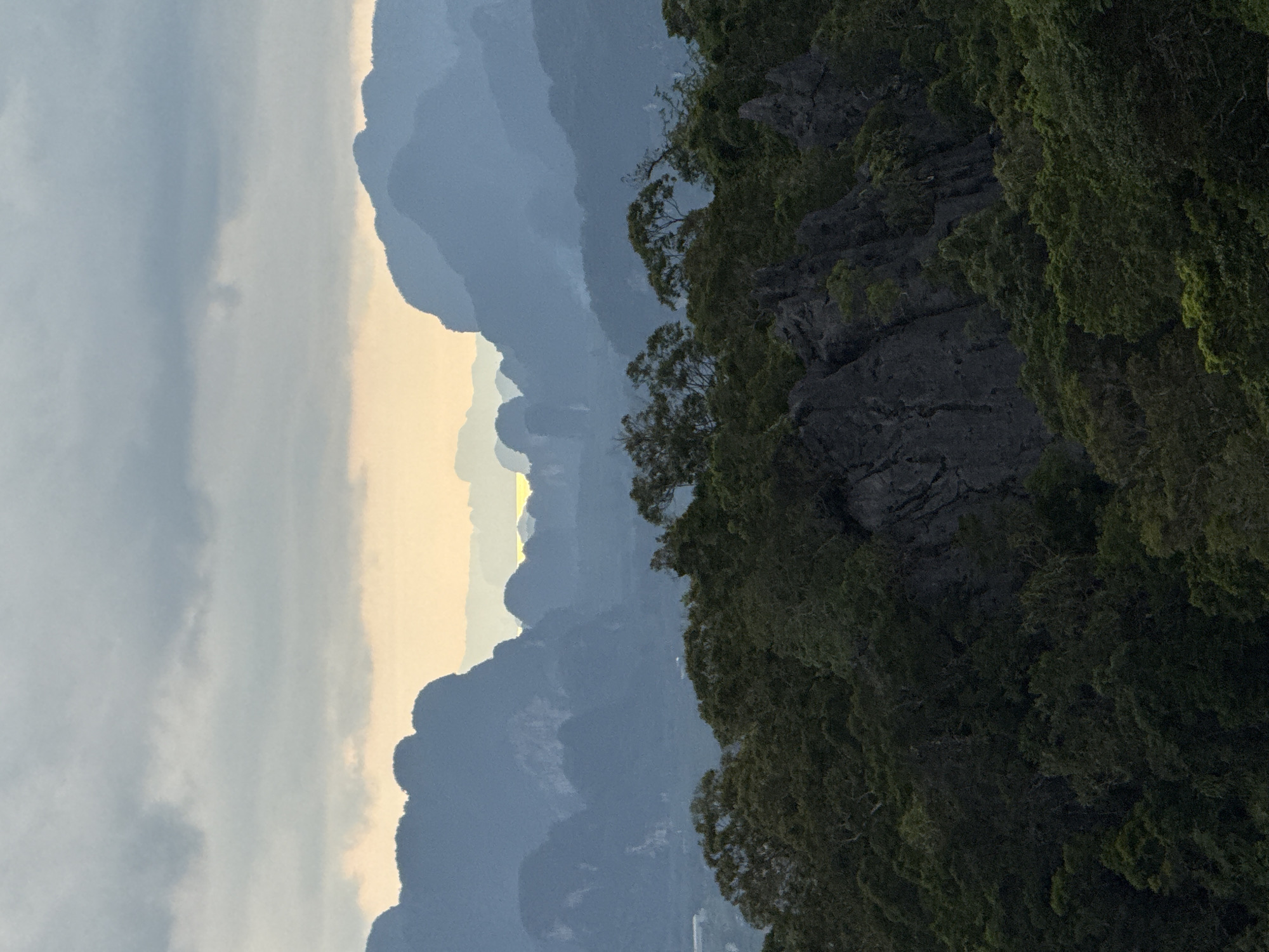 The image depicts a panoramic view of a mountainous landscape at dusk, with layers of mist obscuring the distant peaks. The foreground is dominated by a dense forest of lush green trees and dark, jagged rock formations. The middle ground features more forested hills, and the background consists of receding mountain ranges fading into the hazy horizon. A sliver of pale yellow sunlight is visible in the distance, peeking through a gap in the mountains, suggesting a sunset or sunrise. The sky above is a mosaic of muted gray and blue clouds. The location is identified as Ban Khao Tang, Thailand, a region known for its karst topography and tropical forests.