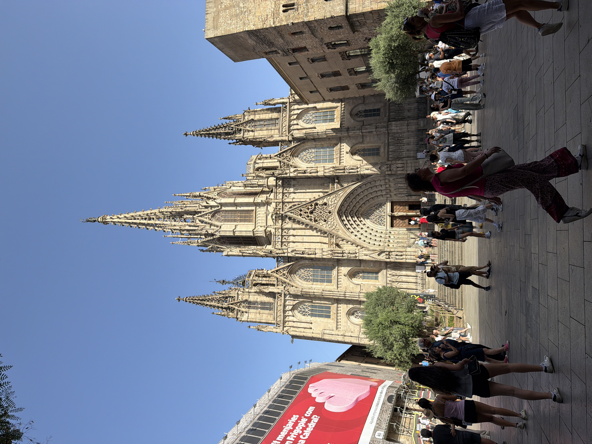 This is an eye-level, outdoor, full-shot photograph of the Barcelona Cathedral in Barcelona, Spain, on a clear, sunny day. The cathedral, a Gothic-style building with intricate stone carvings and tall spires, dominates the center of the frame. The facade features a large, arched entranceway and numerous windows with stained glass. To the left of the cathedral, a large red billboard with white text and a pink heart graphic is visible, partially obscuring a construction site. The foreground is a paved plaza filled with people walking in various directions. The people are dressed in casual summer attire, and many appear to be tourists. Some individuals are walking alone, while others are in groups, engaged in conversation or taking photographs. Trees are scattered throughout the scene, and the sky is a clear blue. The overall atmosphere suggests a busy tourist area on a warm day.