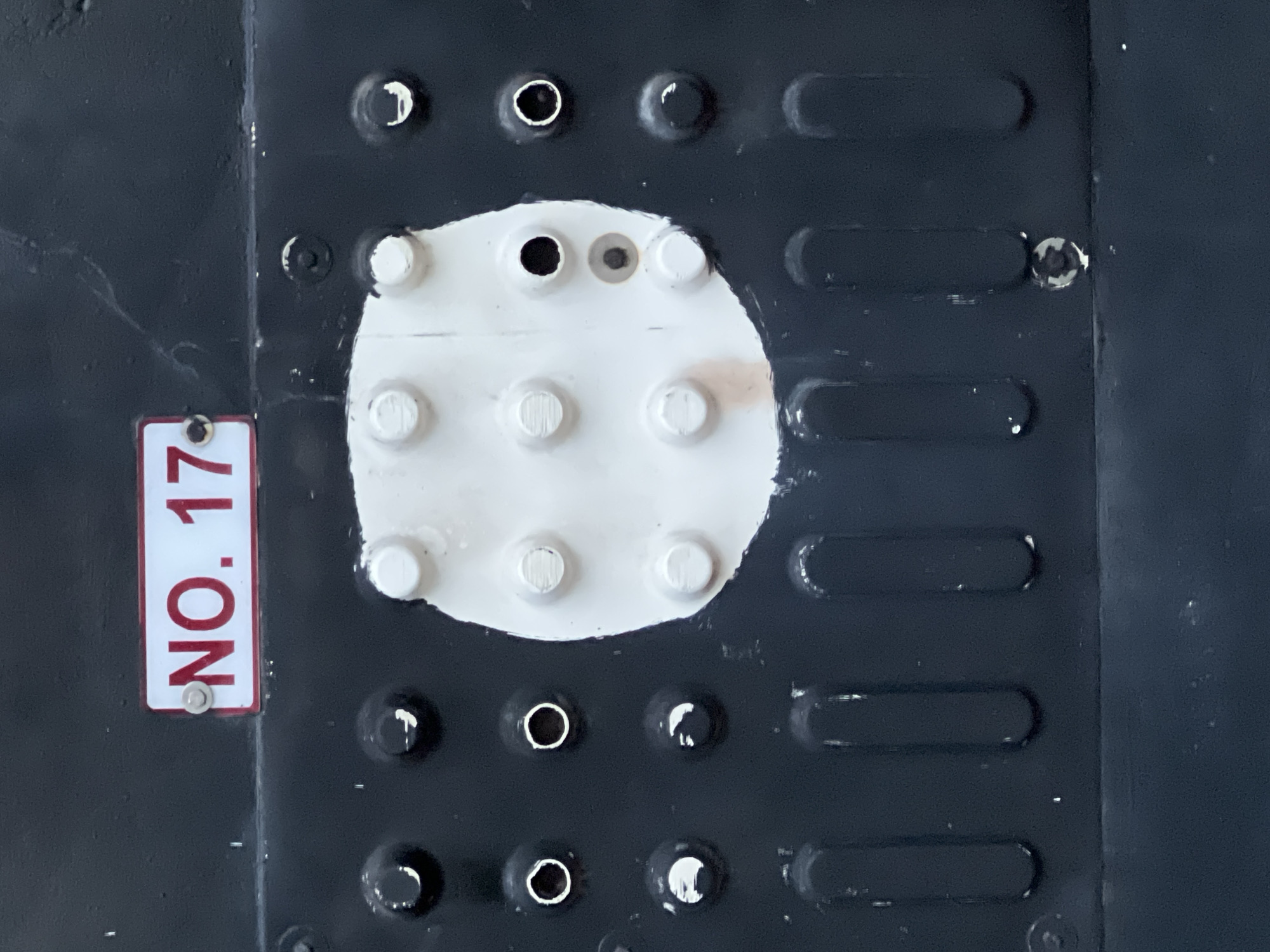 The image shows a close-up, overhead view of a dark, textured surface with a white, roughly circular patch in the center. The white patch has several raised, circular bumps, resembling a keypad or a tactile indicator. Surrounding the white patch, the dark surface has a pattern of small, circular indentations, some with a reflective quality, suggesting they might be holes or recessed buttons.
To the left of the dark surface, a small, rectangular sign with white text on a red background is attached. The text reads "NO. 17". The sign is secured by a small silver screw.
The overall impression is of a functional or industrial surface, possibly a control panel or a textured grip area. The lighting appears to be direct, creating some harsh reflections and shadows on the textured elements. There are no people or explicit signs of activity, but the presence of the numbered sign implies a designated area or object. The setting is unclear, but the robust nature of the surface suggests an outdoor or industrial environment.
