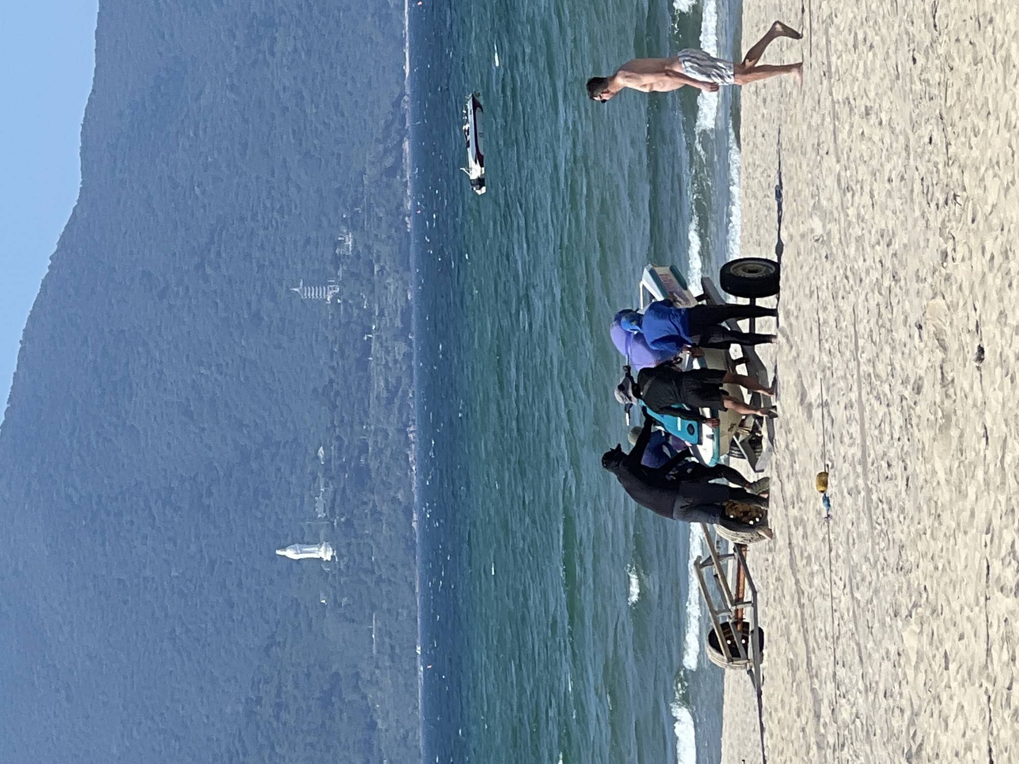 On a sandy beach in Da Nang, Vietnam, a group of four men are working to move a jet ski on a wheeled trailer towards the ocean. The water is a deep blue-green, with moderate waves breaking near the shore. In the background, a lush green mountainside rises, dotted with two structures: a large white statue of a woman and a pagoda. A white and red speedboat is visible on the water in the middle distance. Another man stands on the beach, observing the activity. The sky is clear and blue.