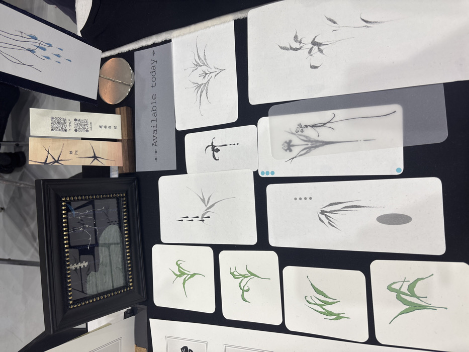 The scene depicts an overhead view of an art display table located in the city of Ban Rong Ruea Kham, Thailand. Various art pieces, primarily minimalist illustrations, are arranged on a dark, flat surface.
In the upper left, a black rectangular frame with gold bead accents encloses a reflective surface with a white and blue abstract design. To its right, two tall, narrow rectangular items, possibly bookmarks or art prints, stand upright in a small wooden holder. The left item features dark, stylized branch-like patterns and Thai script. The right item includes a QR code, similar branch patterns, and Thai script, with visible text "red ochre" and "อวยพร". A small, polished, circular metallic object rests to the right of these items.
Centrally positioned, a grey rectangular sign displays the text "--- Available today ---".
The majority of the table space is covered with white rectangular cards, each with rounded corners, featuring minimalist ink illustrations.
* The leftmost column contains four cards depicting green plant forms, possibly grasses or leaves.
* The center column displays three cards: the top shows a grey bamboo branch with three black teardrop shapes; the middle shows a black orchid-like flower with a single drip; the bottom shows grey bamboo leaves with four small black dots and a grey oval.
* The rightmost section includes two white cards with grey plant illustrations: one symmetrical, the other a multi-stemmed plant. Partially overlaying another white card in the bottom right is a translucent bookmark-like object featuring two slender, dark line-drawn flowers and two light blue dots on its upper left edge.
The background, blurred, reveals a segment of a person's lower legs and dark shoes, standing on a light-colored floor, suggesting an indoor setting.