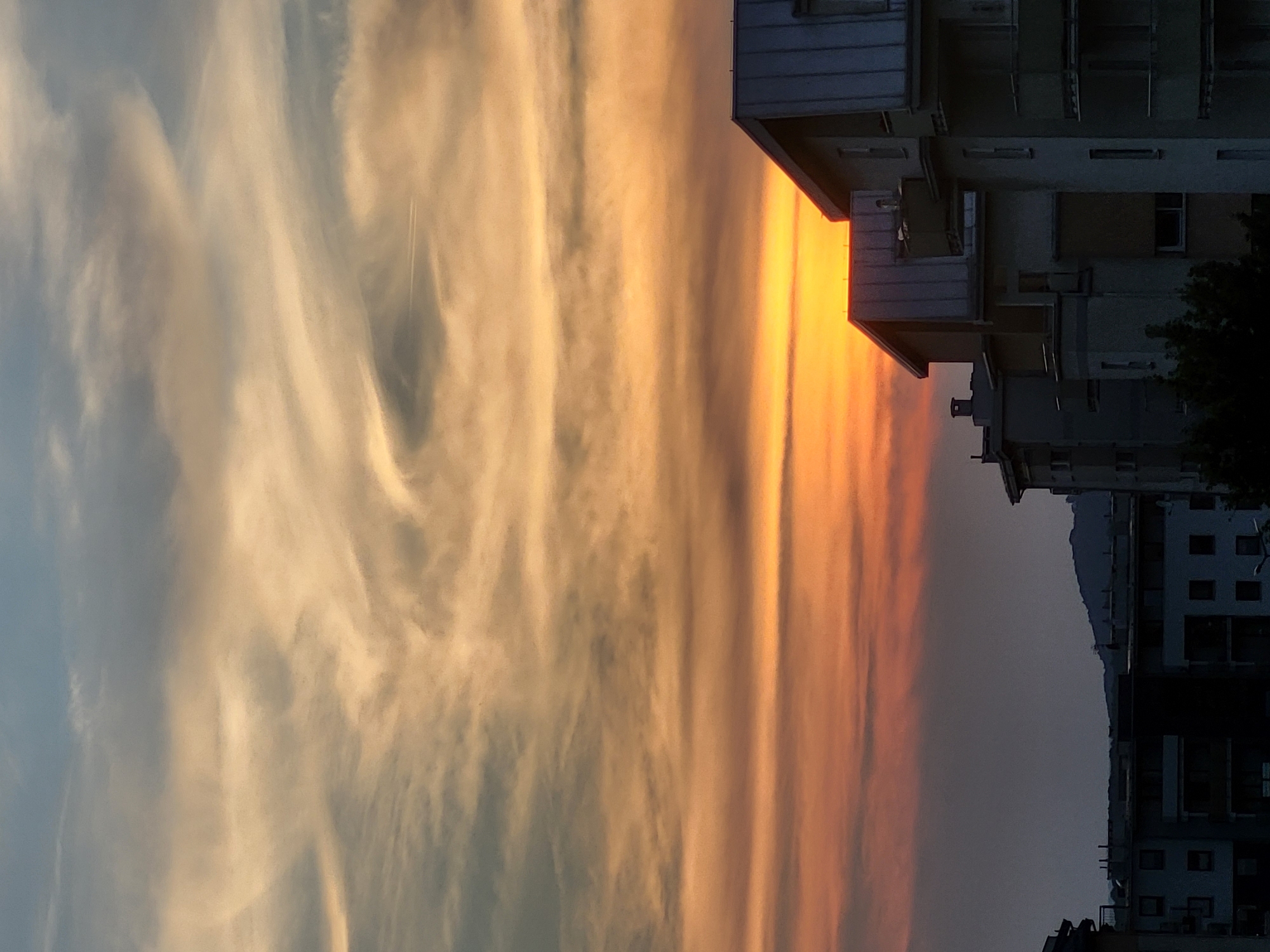 The image depicts a sunset scene over a city. In the foreground, the rooftops and upper stories of apartment buildings are visible, with their grey, sloped roofs and balconies. The buildings are a muted grey and beige color. Behind the buildings, a vibrant sunset fills the sky with bands of orange, yellow, and pink, transitioning to a dusky blue at the top. Wispy clouds are scattered across the sky, with a faint contrail from an airplane visible in the upper portion. In the distance, the silhouettes of hills are visible against the fading light. The overall impression is of an urban landscape at dusk, with the dramatic colors of the sunset providing a striking contrast to the muted tones of the architecture.