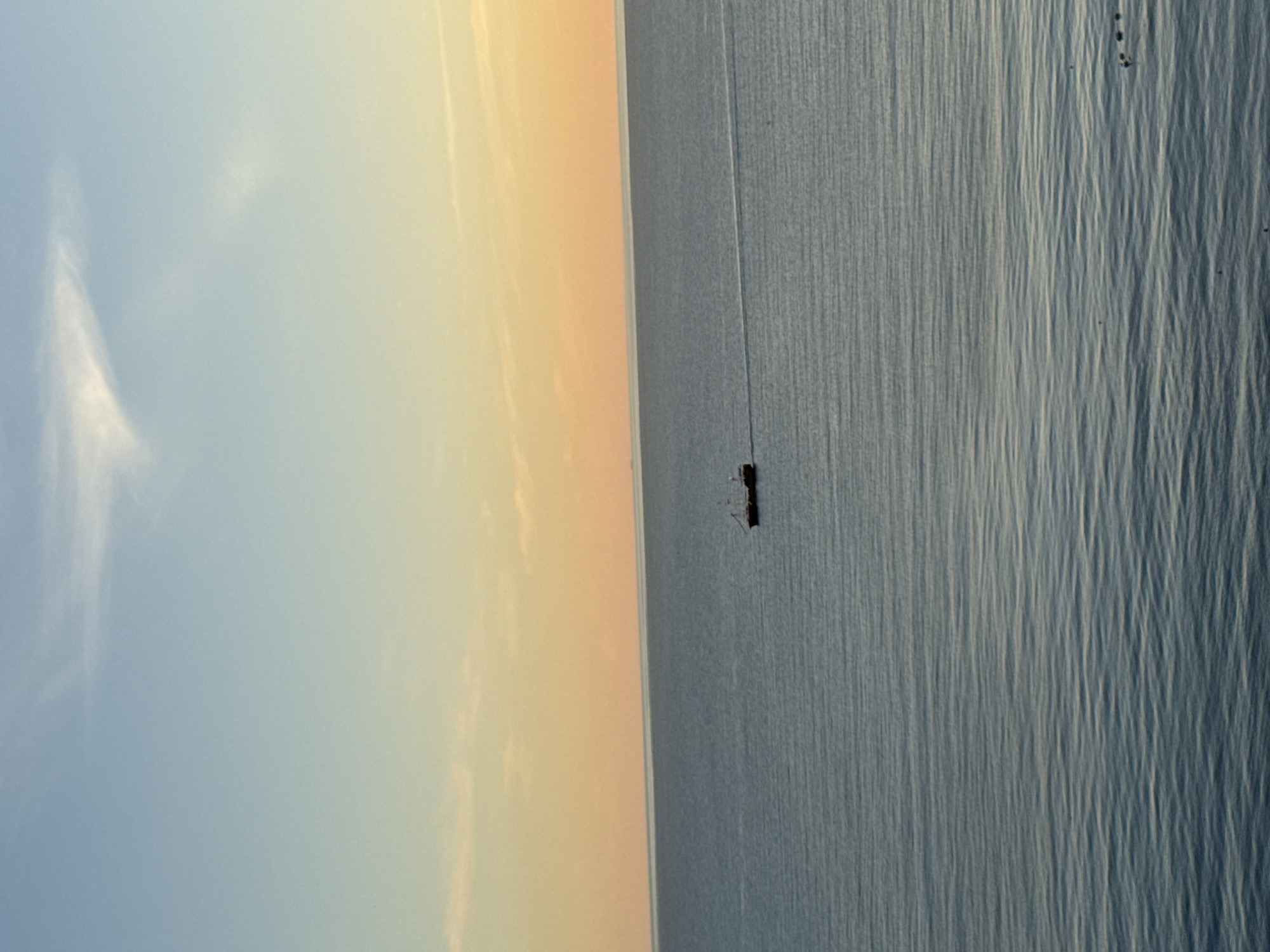 An aerial view captures a vast expanse of calm, deep blue water with subtle ripples under a gradient sky transitioning from pale blue at the top to a warm orange near the horizon. A solitary, dark-hulled vessel, appearing to be a small ship or a large boat with visible masts and rigging, is positioned in the mid-right section of the frame, creating a faint white wake behind it as it travels across the water. The overall scene conveys a sense of tranquility and open space, with no discernible human presence or urban elements visible within the frame. The location is identified as Varvarovka, Russia.