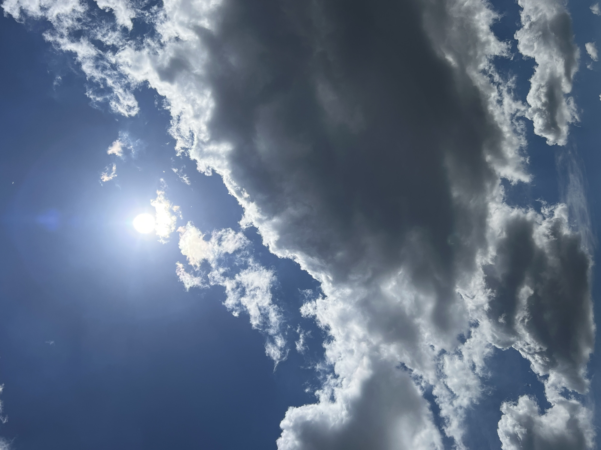 The image depicts a bright, daytime scene with a clear blue sky partially obscured by clouds. The sun is visible in the upper left quadrant, shining brightly with a noticeable lens flare. The clouds range from fluffy white to dark gray, indicating varying thicknesses and potentially different types of cloud formations. The image is taken from a low angle, looking up at the sky. No people, subjects, or objects besides clouds are visible. The location, Vertelka, Ukraine, is not visually verifiable from the image.