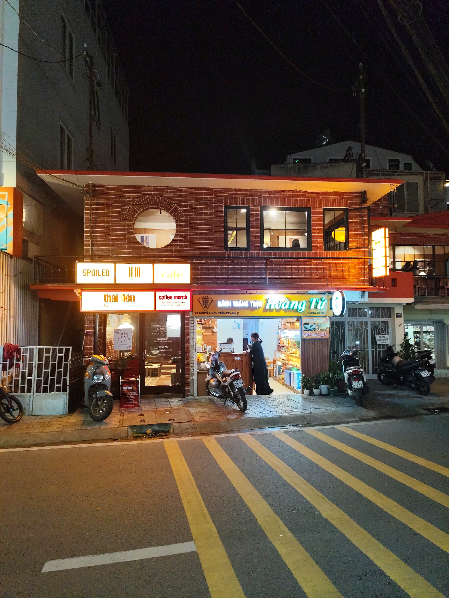 The image depicts a street scene at night in Da Lat, Vietnam. Two businesses are visible: "SPOILED cafe" and "BÁNH TRÁNG THẬT Hoàng Tử." A person is standing outside the "BÁNH TRÁNG THẬT Hoàng Tử" bakery/store, likely interacting with the staff inside. Several motorbikes are parked on the sidewalk near both establishments. The street has yellow and gray crosswalk lines. The address "79 NGUYỄN VĂN TRỐI-P2- ĐÀ LẠT" is visible on a sign, indicating the location within Da Lat. The building architecture features a brick facade with a round window on the second level above "SPOILED cafe."