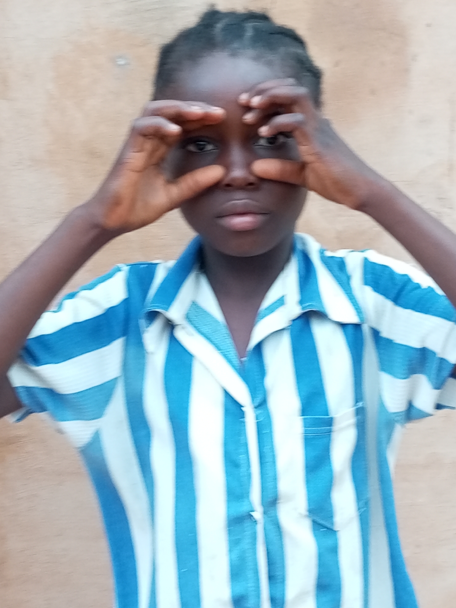The image shows a medium shot of a young girl in Tempure, Nigeria. She is wearing a blue and white striped shirt and holding her hands up to her eyes, as if looking through binoculars. The background is a plain, light brown wall. The lighting is natural and appears to be outdoors. There is no other visible activity or interaction in the scene.