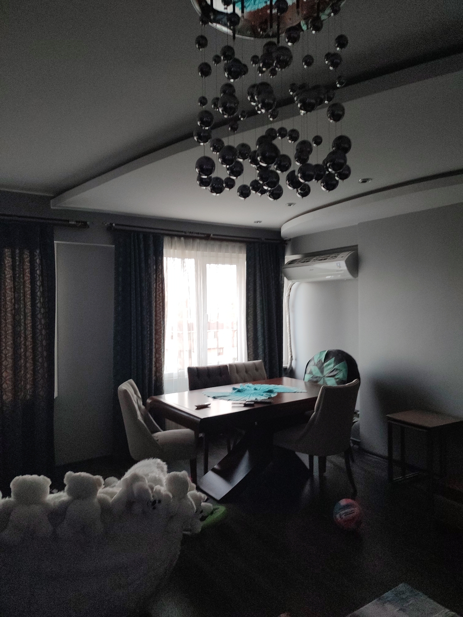This image depicts a dimly lit dining room, likely in an apartment in Kartal, Turkey, given the context. The room features a dark wooden dining table with several chairs around it. A large window with white sheers is visible, covered by dark curtains on either side. Above the dining table, a modern, abstract chandelier with numerous dark, spherical elements hangs from the ceiling. An air conditioning unit is mounted on the wall to the right of the window. In the foreground, a collection of white stuffed animals, possibly teddy bears, are arranged on what appears to be a fuzzy white rug or blanket. A colorful ball is on the dark wooden floor near the dining table. The overall ambiance is subdued, with minimal natural light entering the room.