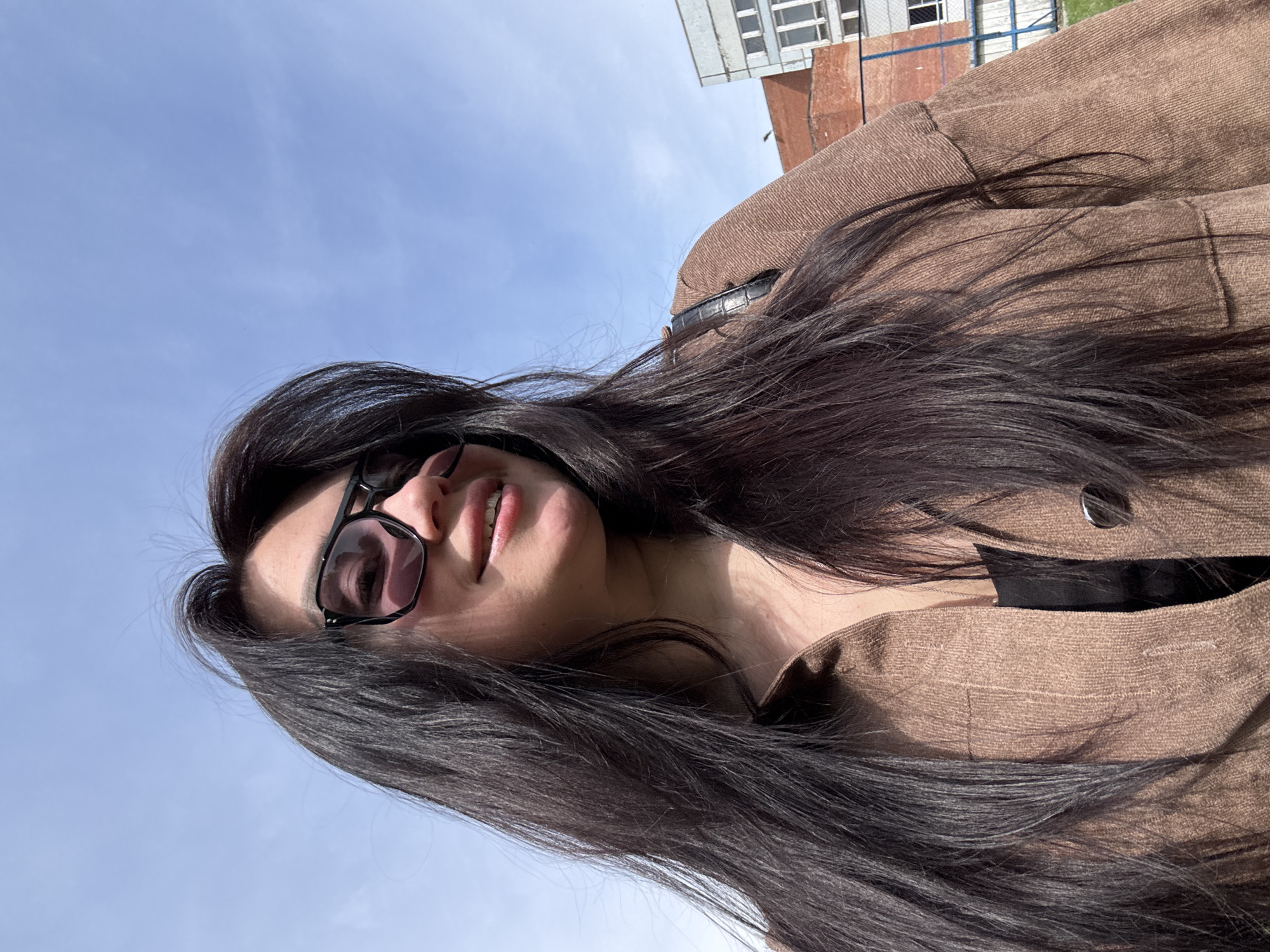 A woman with long, dark hair and sunglasses looks up into the camera under a bright blue sky. She wears a brown jacket with visible button and black trim around the neckline. In the blurred background, a building with white window frames and a brick structure are visible. The location appears to be outside in an urban environment. The location is in the city of Unknown, Unknown.