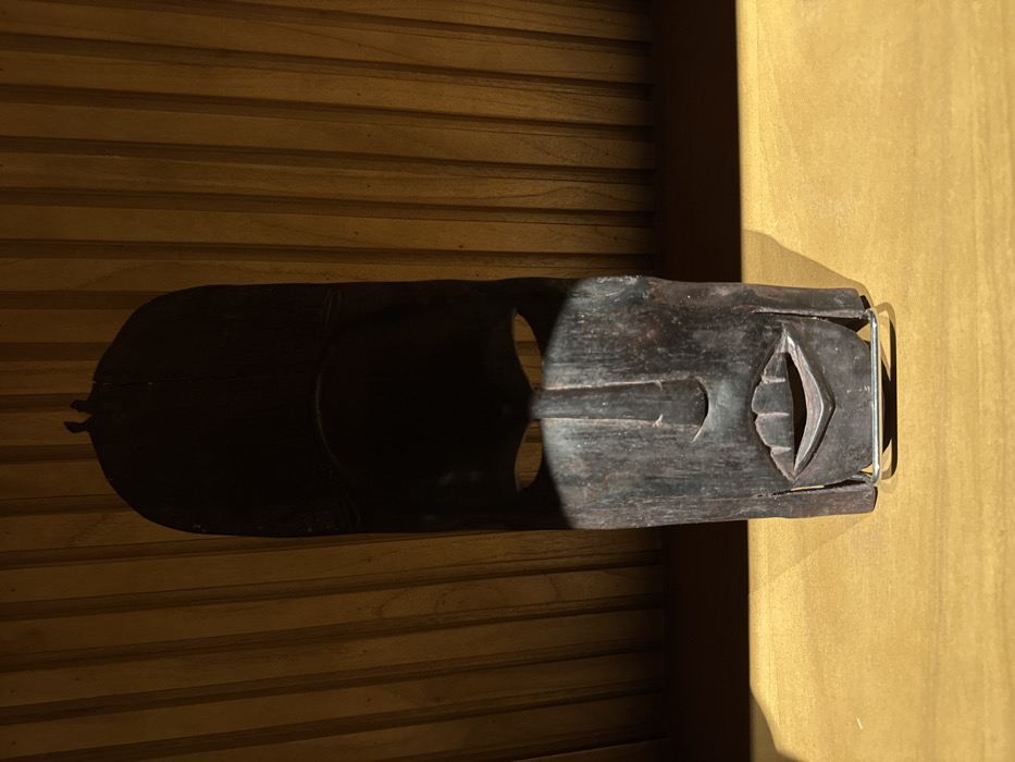 A wooden mask is positioned on a horizontal surface featuring parallel lines. The mask, dark in color, displays stylized facial features, including eye sockets and a mouth. A metal wire or bracket is visible at the mask's edge. Light casts a shadow that extends across the textured surface, creating a contrast with a brighter area on the adjacent, smoother surface.