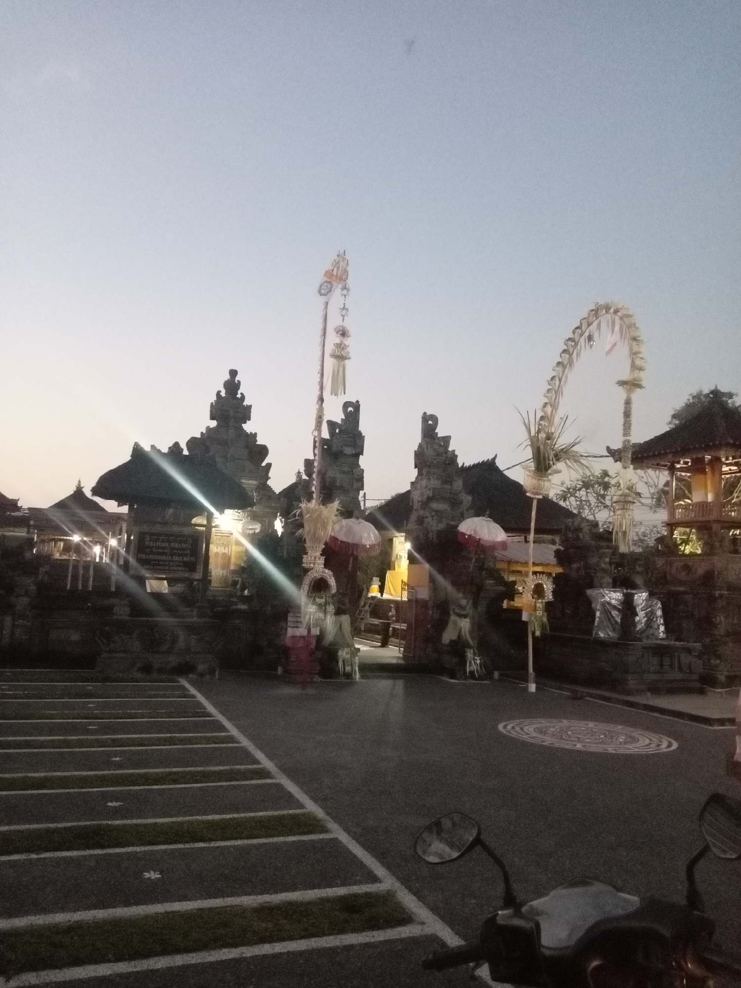 A wide shot of an outdoor temple complex at dusk in an unknown location. The sky is a pale grey, transitioning to a lighter hue near the horizon. The temple features traditional Balinese architecture with intricate stone carvings and tiered roofs. Several tall, ornate poles with decorative elements are placed strategically, some adorned with streamers and foliage. White umbrellas are also visible, possibly as part of a ceremony or decoration. In the foreground, a parking lot is delineated with white lines and patches of grass. A motorcycle is parked in the lower right corner of the frame. The overall atmosphere is tranquil and dimly lit, suggesting the end of a day or the beginning of an evening.