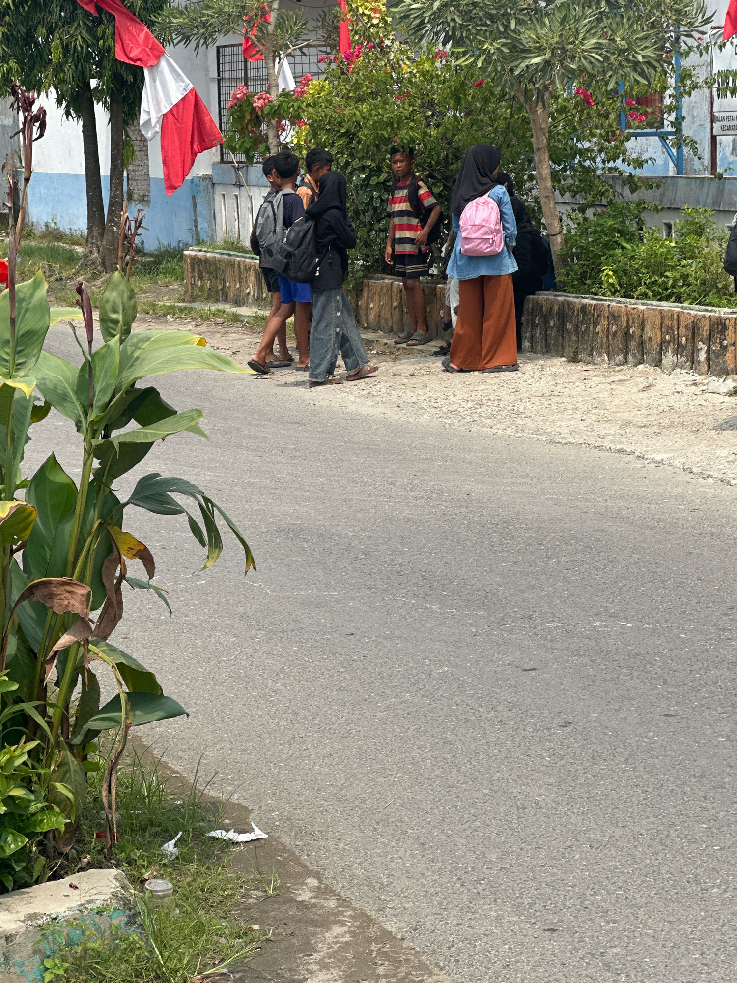 In the City of Binjai, Indonesia, a group of four young people are standing on a paved walkway next to a road. Three of them are boys, and one is a girl wearing a hijab. The boys are dressed casually, with one wearing a striped t-shirt and shorts, another in a blue shirt and shorts, and the third in a dark shirt and pants. The girl is wearing a denim shirt and brown pants. All four have backpacks on. A red and white Indonesian flag is visible hanging in the background, suggesting a celebratory or patriotic occasion. The environment is somewhat overgrown with vegetation, with a large green plant in the foreground. The walkway is bordered by a low, weathered wall. The scene appears to be outdoors on a clear day.