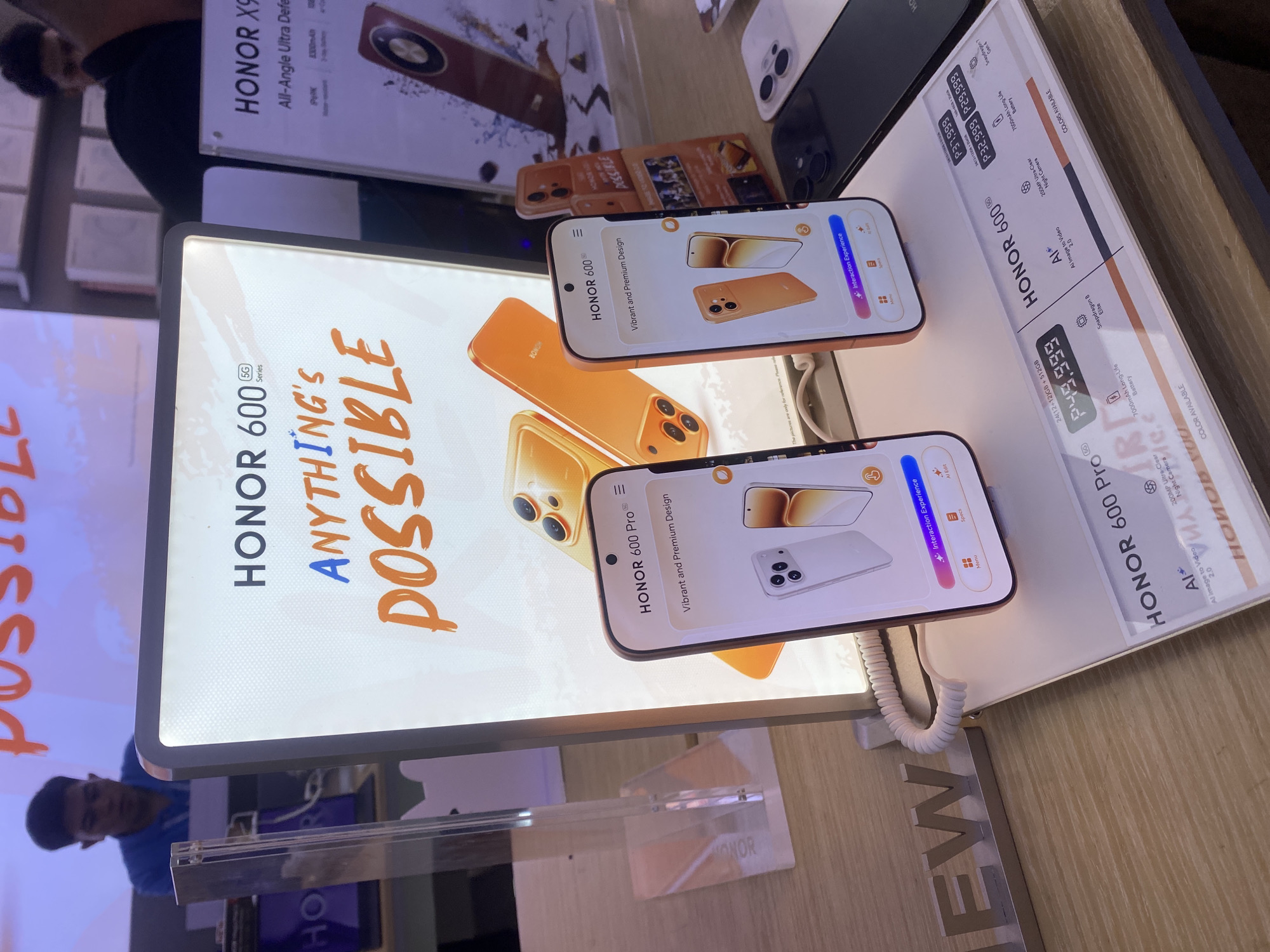 The image depicts a retail display of Honor smartphones in a store, likely in Davao City, Philippines, as indicated by the currency format "P49,999" and "P37,999". The main focus is on the Honor 60 Pro and Honor X9 models. Two Honor 60 Pro phones are prominently displayed, with one showcasing a vibrant screen displaying "Vibrant and Premium Design" and offering an "Interaction Experience" feature. The other Honor 60 Pro phone is positioned on a stand next to the display. In the background, a large illuminated sign features the text "HONOR 60 Series ANYTHING'S POSSIBLE" with the word "POSSIBLE" in an orange, brushstroke font. To the right, a display for the Honor X9 is partially visible, showing its specifications and an image of the phone. A person, likely a store employee or customer, is partially visible in the blurred background behind the main display, wearing a blue shirt. The overall environment suggests a modern electronics retail store with wooden shelving and bright lighting.