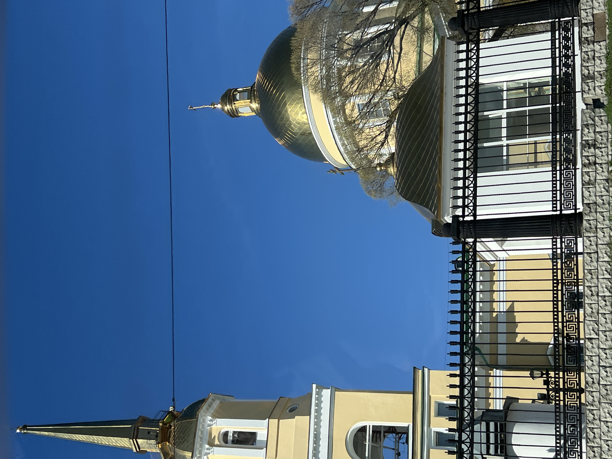 This image captures a bright, sunlit view of a Russian Orthodox church complex in Izhevsk, Russia, set against a clear, deep blue sky. The scene is dominated by two distinct church structures, both adorned with radiant golden domes and spires.
On the right, a building features a large, spherical golden dome, crowned with a cross at its apex. The dome's surface reflects the sunlight, creating bright highlights. Partially obscuring the dome and the light yellow facade of the building are numerous bare tree branches, suggesting either early spring or late autumn. Architectural details in white trim are visible on the building's exterior.
To the left, a tall, slender golden spire, also topped with a cross, rises prominently. Below it is a smaller, golden onion-shaped dome. This structure appears to be a bell tower or another section of the same church complex, sharing the same light yellow color palette with white decorative elements. Thin, dark utility wires or cables are visible stretching across the sky, connecting near the top of this spire.
In the foreground, a detailed black wrought-iron fence acts as a boundary. The fence features ornate vertical bars and horizontal sections adorned with a repeating Greek key pattern.
The lighting indicates a clear, sunny day, likely midday, with strong contrasts and reflections on the golden surfaces. There are no people visible, and no specific activity is taking place. No discernible text is present in the image.