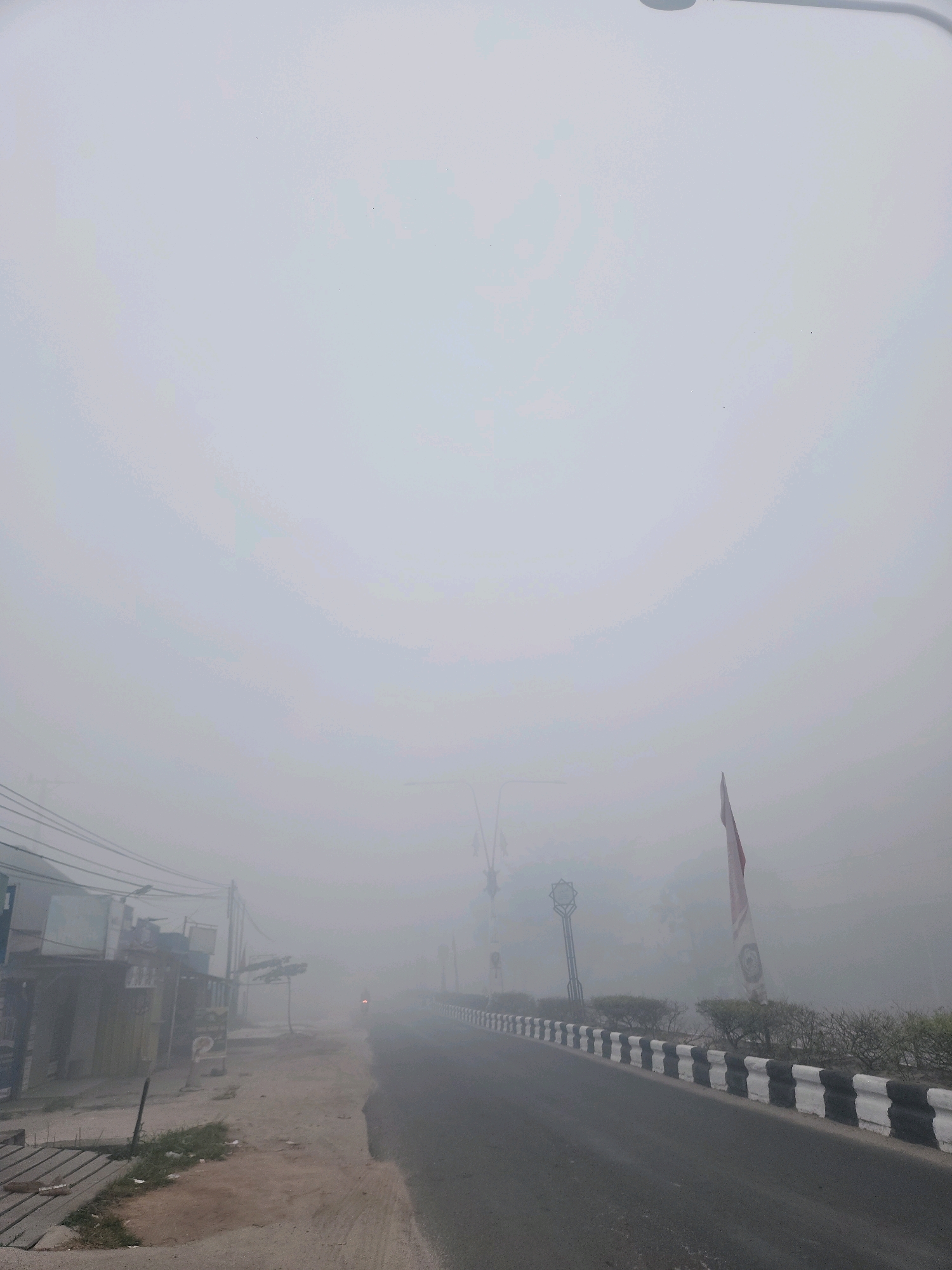 The image depicts a street scene in Baamang Tengah, Indonesia, enveloped in a dense, white fog. The fog obscures visibility significantly, creating a hazy and diffused atmosphere. A paved road runs diagonally from the bottom right towards the center of the image. On the left side of the road, there are several buildings with visible signage, some appearing to be small shops or residences. Electrical wires and poles are also present. A dirt path runs alongside the buildings. On the right side of the road, a barrier with black and white stripes separates the road from a grassy area with sparse vegetation and trees. A flag, predominantly red and white, is visible on the right side, and a lamppost with a decorative top is situated in the middle ground, also on the right. A faint red light, possibly from a vehicle, is visible in the distance on the road, indicating minimal traffic. The overall impression is one of a quiet, obscured morning in a semi-urban or rural setting, with the fog being the dominant environmental factor.