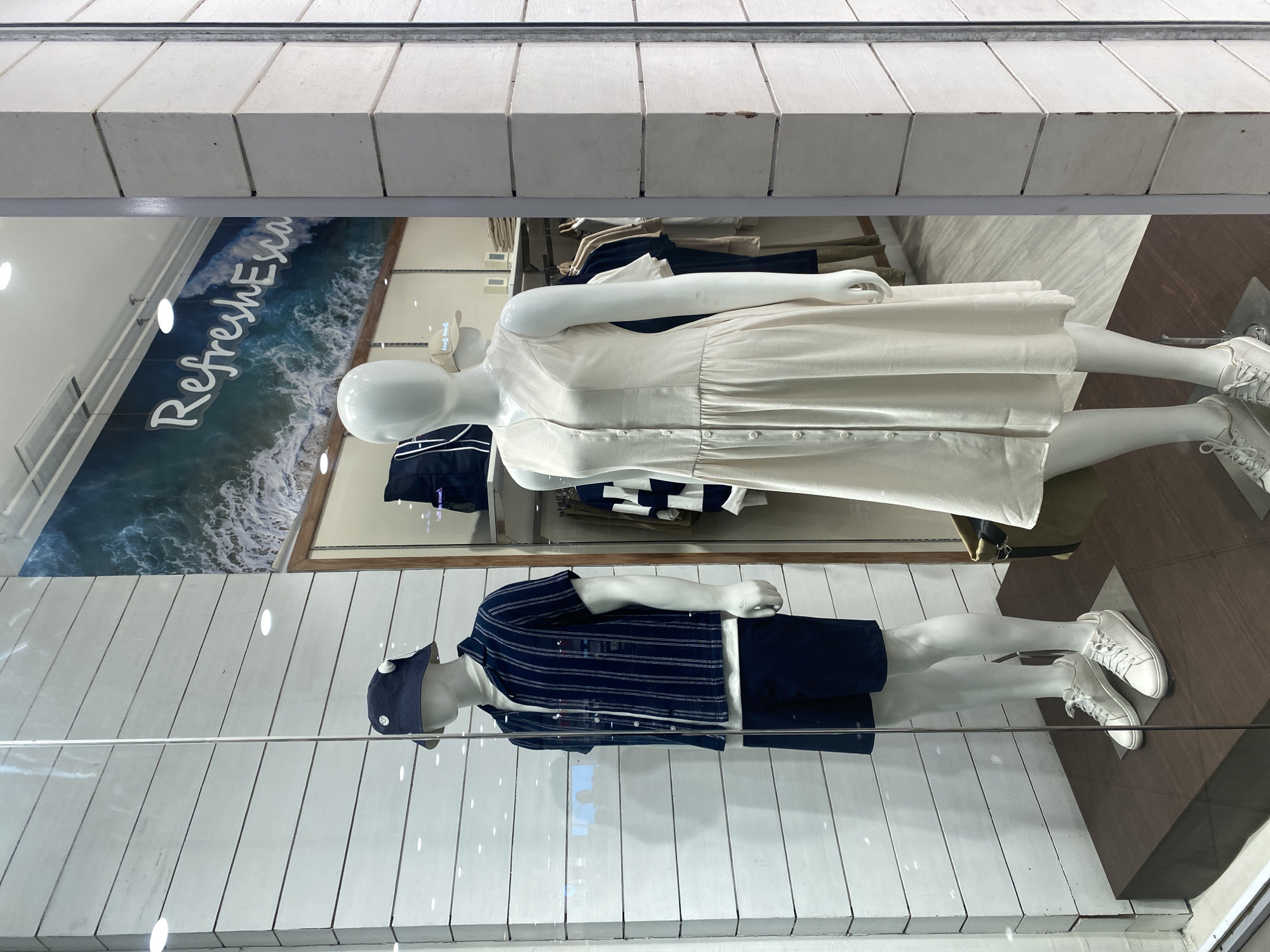 The provided image displays a retail shop window in Davao City, Philippines, showcasing two mannequins modeling casual apparel. The scene is viewed through a reflective glass barrier, with subtle reflections of overhead lighting visible across the surface.
On the right side of the display, a female mannequin is dressed in a flowing, cream or off-white button-down dress with a gathered waist, paired with white lace-up sneakers. Her pose is natural, with one arm gently bent.
On the left, a male mannequin wears a dark blue short-sleeved shirt featuring thin white horizontal stripes, dark blue shorts, and a matching dark blue cap. He is also adorned with white lace-up sneakers.
The background of the display features a large mural depicting a dynamic ocean scene with breaking waves, evoking a sense of the seaside. Overlaid on this ocean graphic, in a stylized white script, is the partially visible text "RefreshEsca," suggesting a theme related to "refreshment" or "escape." The display floor beneath the mannequins is segmented, with a lighter, possibly white, wood-paneled section on the left and a darker, polished brown section on the right. Above the display, a textured, light-colored architectural element with horizontal segments is visible, possibly part of the building's facade or interior ceiling. The overall impression is a brightly lit, summer-themed fashion presentation, consistent with coastal living.