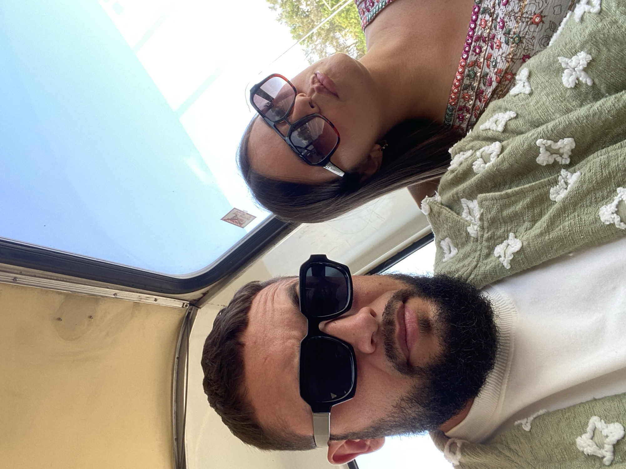 In an outdoor setting, a man and a woman are captured in a selfie taken from inside a vehicle, possibly a tuk-tuk, given the interior view of the ceiling and the open window. The man, in the foreground on the left, wears black-rimmed sunglasses and a light green jacket adorned with white floral appliques over a white sweatshirt. His dark hair is short, and he has a beard. The woman, on the right, is partially visible, also wearing sunglasses with a dark frame and a patterned top featuring floral embroidery. The background shows a bright blue sky and some greenery, suggesting a sunny day. A small red and white sign is attached to the vehicle's window frame. The lighting suggests it is daytime. There is no specific visual information in the image that allows for an identification of the location within Chaikovsky, Russia.