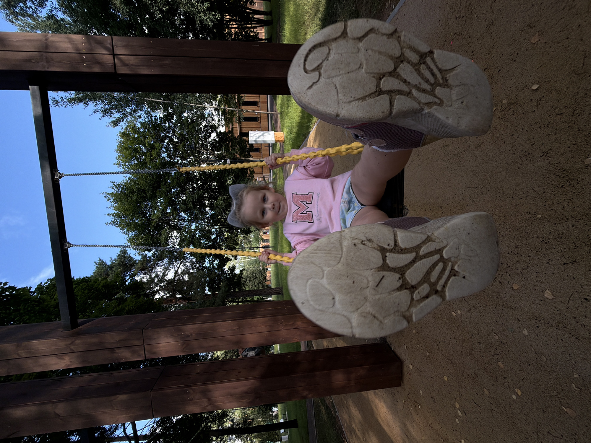 A young girl with blonde hair and cat ear accessories is swinging on a yellow swing. She is wearing a pink long-sleeved shirt with a large "M" logo and light blue denim shorts. The swing is attached to a wooden and metal frame, located outdoors in a park. The ground beneath the swing is covered in sand. Trees and a building are visible in the background. The perspective is from below, looking up at the girl, with her feet and the soles of her shoes prominently featured. The scene appears to be in Malakhovka, Russia, based on the provided context.