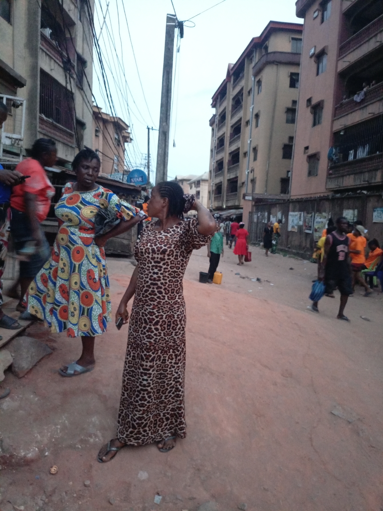 Two women stand on a sloped, dirt-covered street in Onitsha, Nigeria. The woman on the left wears a brightly patterned dress with a blue, yellow, red, and white design. The woman on the right wears a long, leopard-print dress. Both are wearing sandals. Behind them, other people walk along the street. Several multi-story buildings line the street, and power lines are visible overhead. The overall scene is a typical street scene in a densely populated urban area.