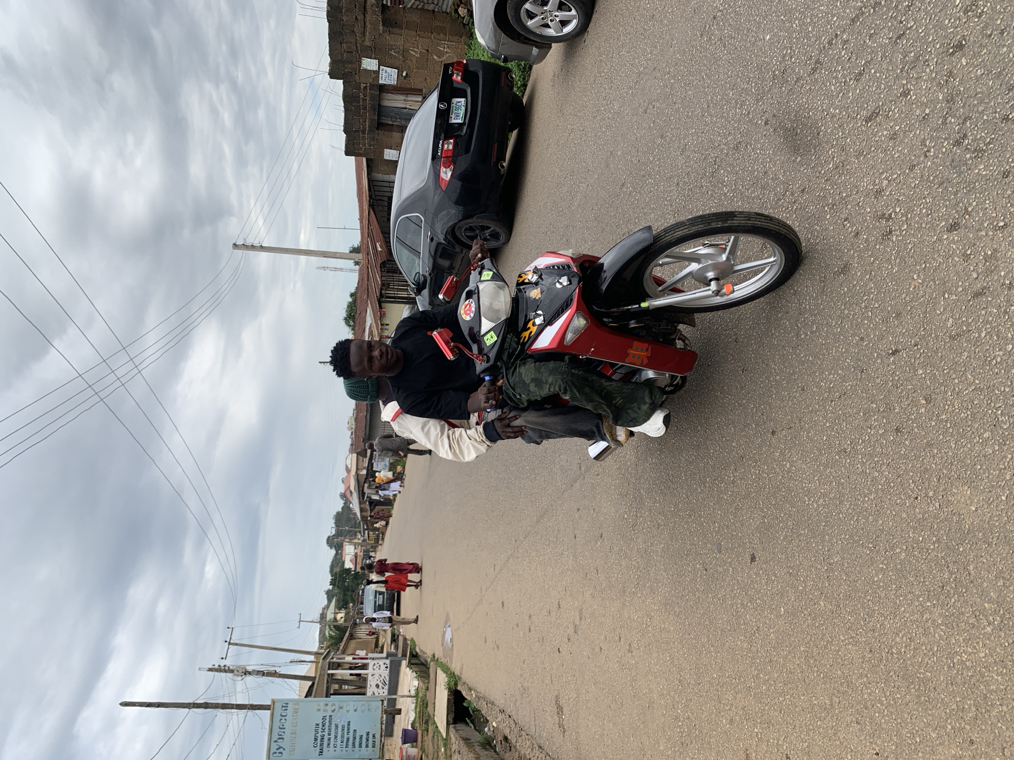 The image depicts a street scene in Jos, Nigeria, on an overcast day. The foreground is dominated by a red and black motorcycle with two young Black men riding it. The rider in front wears a black t-shirt and camouflage pants. The passenger behind him wears a light-colored jacket over a dark garment and a green beanie. Both men appear to be looking forward. The motorcycle has intricate decals, including flame designs and characters on the front panel.
The street itself is paved with asphalt and shows signs of wear, with some patches and debris. To the left of the road, there is a concrete drainage channel and a building with a sign that reads "Cybercom Business Centre." The sign lists various services like "Computer Training School," "Online Registration," "ICT Accessories," "Typing/Printing," and "Lamination." Further down the street on the left, more buildings and stalls are visible, with a few pedestrians walking.
On the right side of the street, a black Acura sedan with a Nigerian license plate (BWR-96GN) is parked. Beside it, another vehicle, partially visible, has its front wheel and fender showing. In the background, the street stretches towards buildings with corrugated iron roofs and some trees. The sky is heavily clouded, suggesting it might rain or has recently rained. The overall atmosphere of the image is that of a typical urban environment in Nigeria.