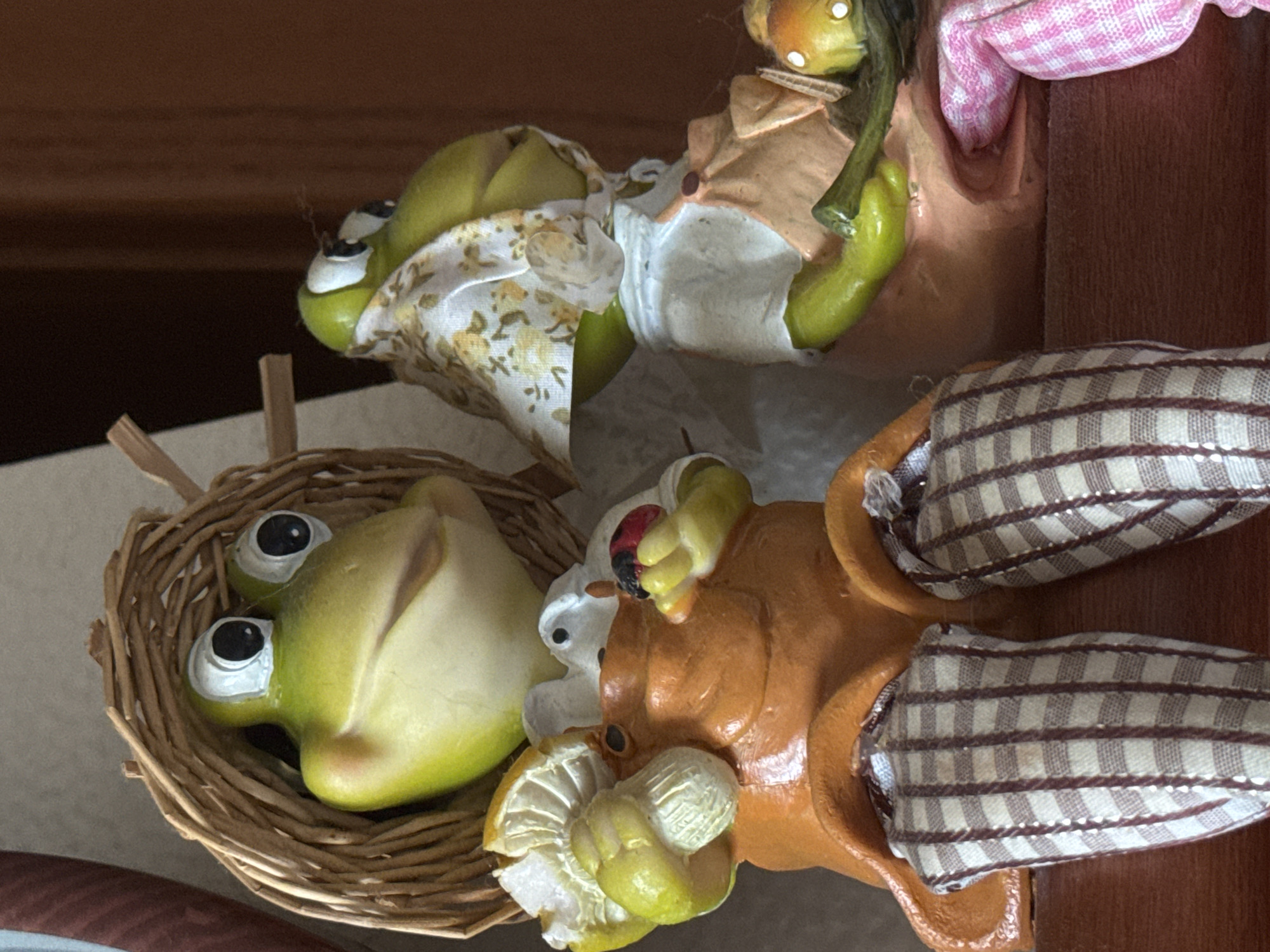 Two ceramic frog figurines are displayed on a wooden surface. The frog on the left is wearing a woven straw hat and an orange jumpsuit with plaid leggings. It is holding a mushroom in its left hand and a ladybug in its right. The frog on the right is wearing a floral headscarf and a light-colored shirt with an orange apron over it, and checkered pink pants. It is holding a green vegetable in its left hand and a mushroom in its right. The background is a blurred wooden surface. The exact location within Kholmsk, Russia, is not discernible from the image.