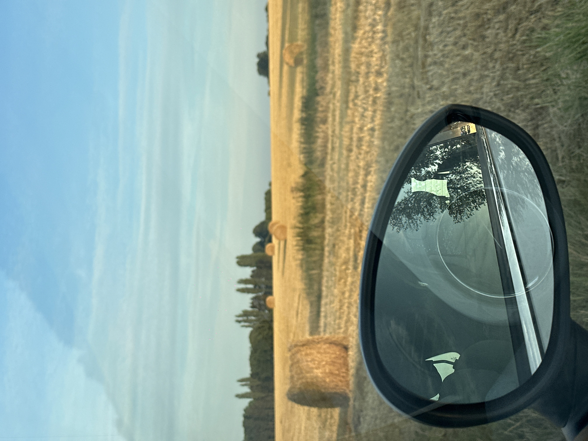 The image depicts a rural scene with hay bales scattered across a harvested field under a blue sky with wispy clouds. In the foreground, a car's side mirror reflects the passing landscape, including trees and the interior of the car, with a silhouette of a person visible. The scene appears to be located in the countryside near Trouville-Alliquerville, France, given the characteristic flat, open fields of the Pays d'Auge region. The lighting suggests it is either early morning or late afternoon, with the sun casting a warm glow on the dry hay. No specific actions or interactions are evident, other than the implied movement of the car capturing the reflection.