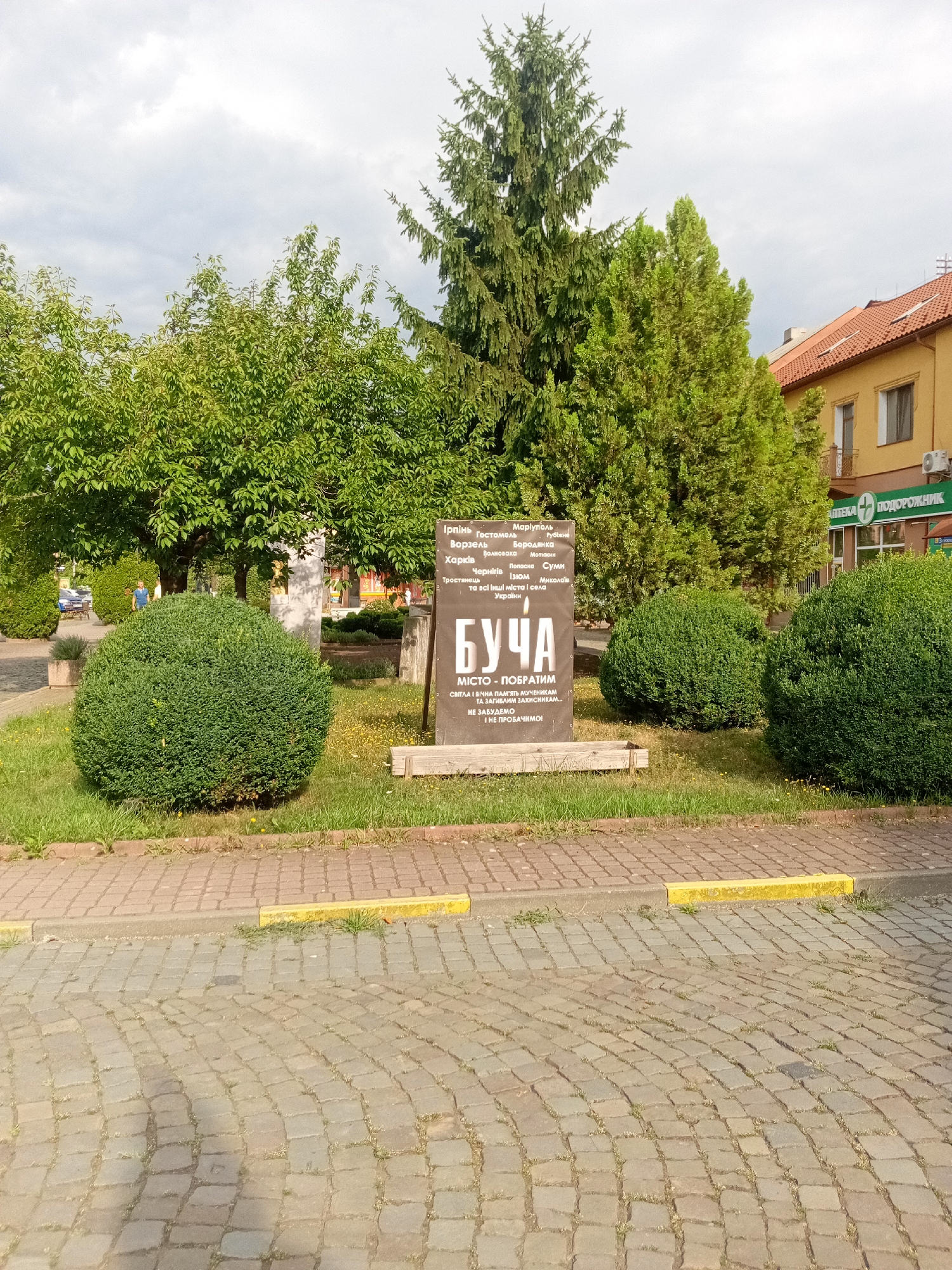 The image depicts a street scene in Tiachiv, Ukraine, featuring a commemorative monument and surrounding greenery. The monument, a dark-colored rectangular plaque mounted on a low wooden base, bears Ukrainian text. The largest text prominently reads "БУЧА", with smaller text below indicating "МІСТО - ПОБРАТИМ" (City - Sister City). Above "БУЧА," a list of other cities is visible, including "Ірпінь," "Маріуполь," "Харків," and "Волноваха." The monument is situated on a patch of grass, bordered by a paved walkway with yellow markings.
In the background, several manicured, spherical green bushes are positioned around the monument. Behind the bushes, lush green trees form a dense canopy, with a tall evergreen tree dominating the right side of the frame. To the far right, a building with an orange-tiled roof and yellow facade is visible, with a green sign on its wall that reads "АПТЕКА ПОДОРОЖНИК" (Pharmacy Podorozhnik) and a green plus sign. A person in a blue shirt is partially visible in the mid-ground, walking near a parked car. The ground in the foreground is paved with cobblestones. The sky is overcast with scattered clouds.