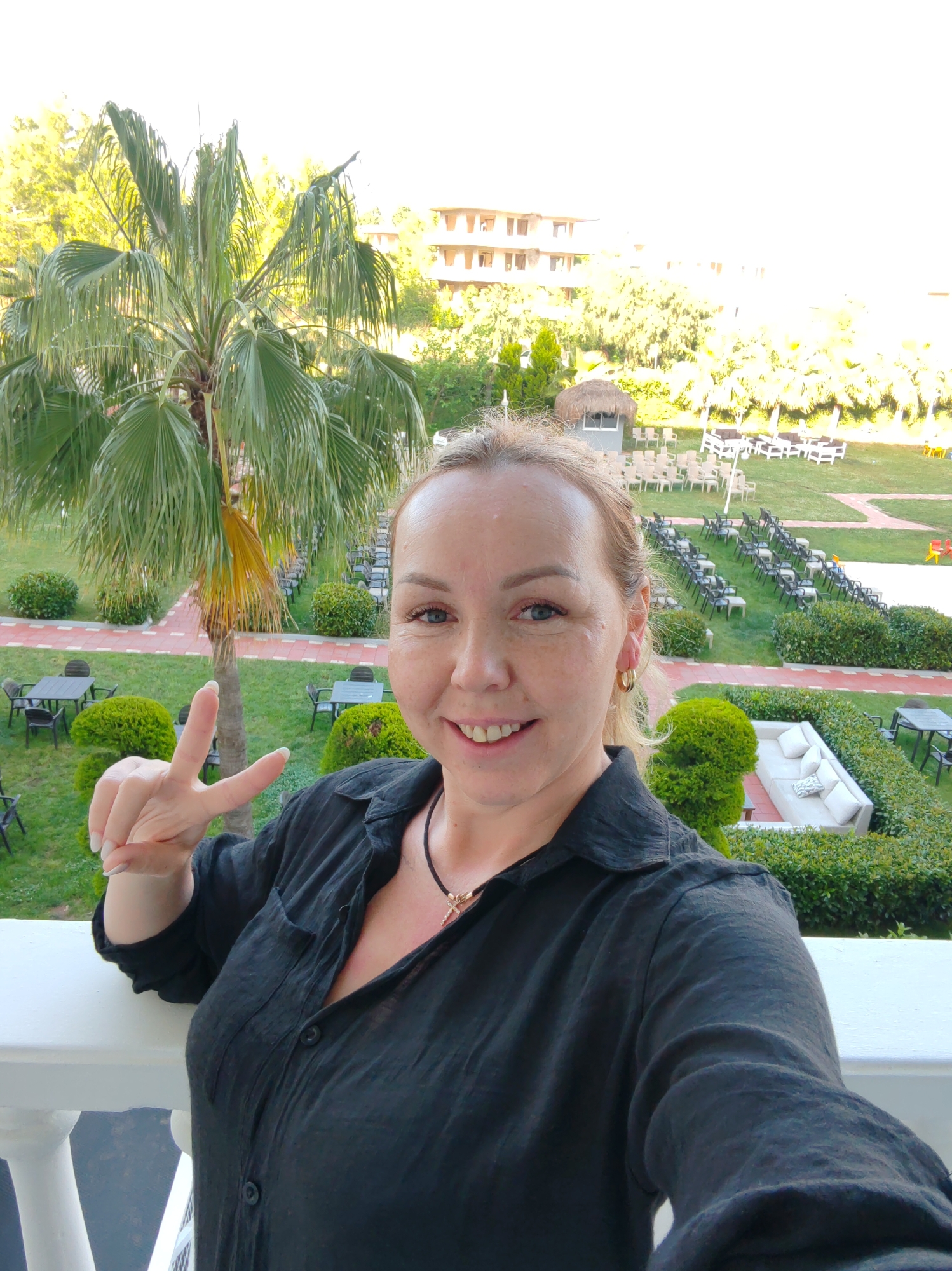 A woman poses for a selfie on a balcony overlooking a resort landscape. She wears a black button-down shirt and gold hoop earrings, holding up two fingers in a peace sign. Beyond the white balcony railing, a palm tree stands to the left, with neatly arranged rows of chairs and tables on a green lawn extending towards a building in the background. A thatched-roof structure is visible among the rows. The scene is bathed in warm sunlight. This location is Alanya, Turkey.