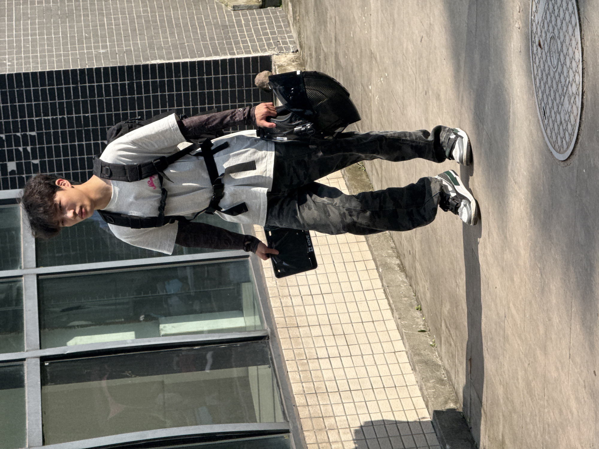 A young man stands outdoors, his body angled toward the viewer. He is wearing a light-colored t-shirt over a dark long-sleeved shirt, paired with camouflage pants and sneakers. A black backpack is strapped to his shoulders, and he is holding a black helmet in his right hand. The man appears to be looking down or to the side, with a neutral expression.
The setting appears to be an urban or institutional environment, characterized by a tiled sidewalk, a building with large glass windows, and a dark grated wall. Sunlight casts distinct shadows, suggesting it is daytime. The weather appears to be sunny. A circular metal manhole cover is visible on the right side of the frame. The scene is captured in Jiangjin, China. No explicit text is visible on the man's clothing, helmet, or surrounding objects. The overall impression is of a casual moment, perhaps before or after an activity involving the helmet, such as riding a motorcycle or bicycle.