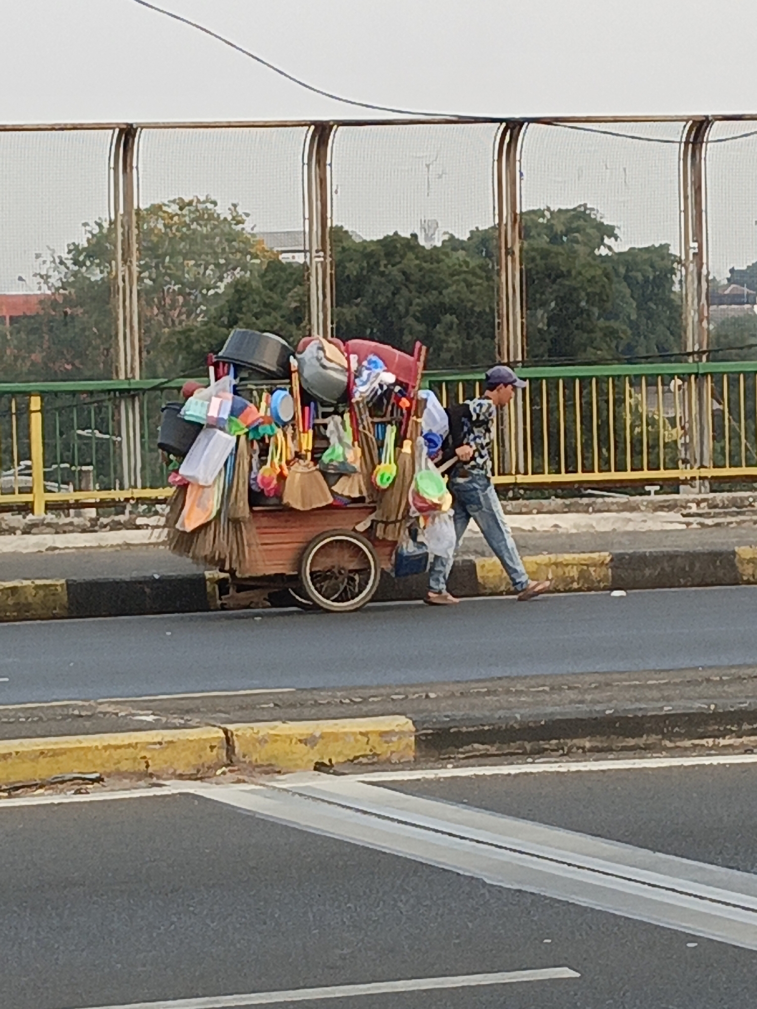In East Jakarta, Indonesia, a man is seen walking alongside a cart loaded with cleaning supplies. The cart, which appears to be old and wooden, is laden with various items including buckets, brooms, dustpans, and what seem to be cleaning cloths or bags. The man, wearing a blue cap, a patterned shirt, and jeans, is pushing or pulling the cart. He has a backpack on his shoulders. The scene takes place on the side of a road, likely a street in the city. In the background, a bridge or overpass with a yellow and green railing is visible, along with trees and some buildings. The road itself has yellow and black markings on the curb. The overall impression is of a street vendor navigating the urban environment.