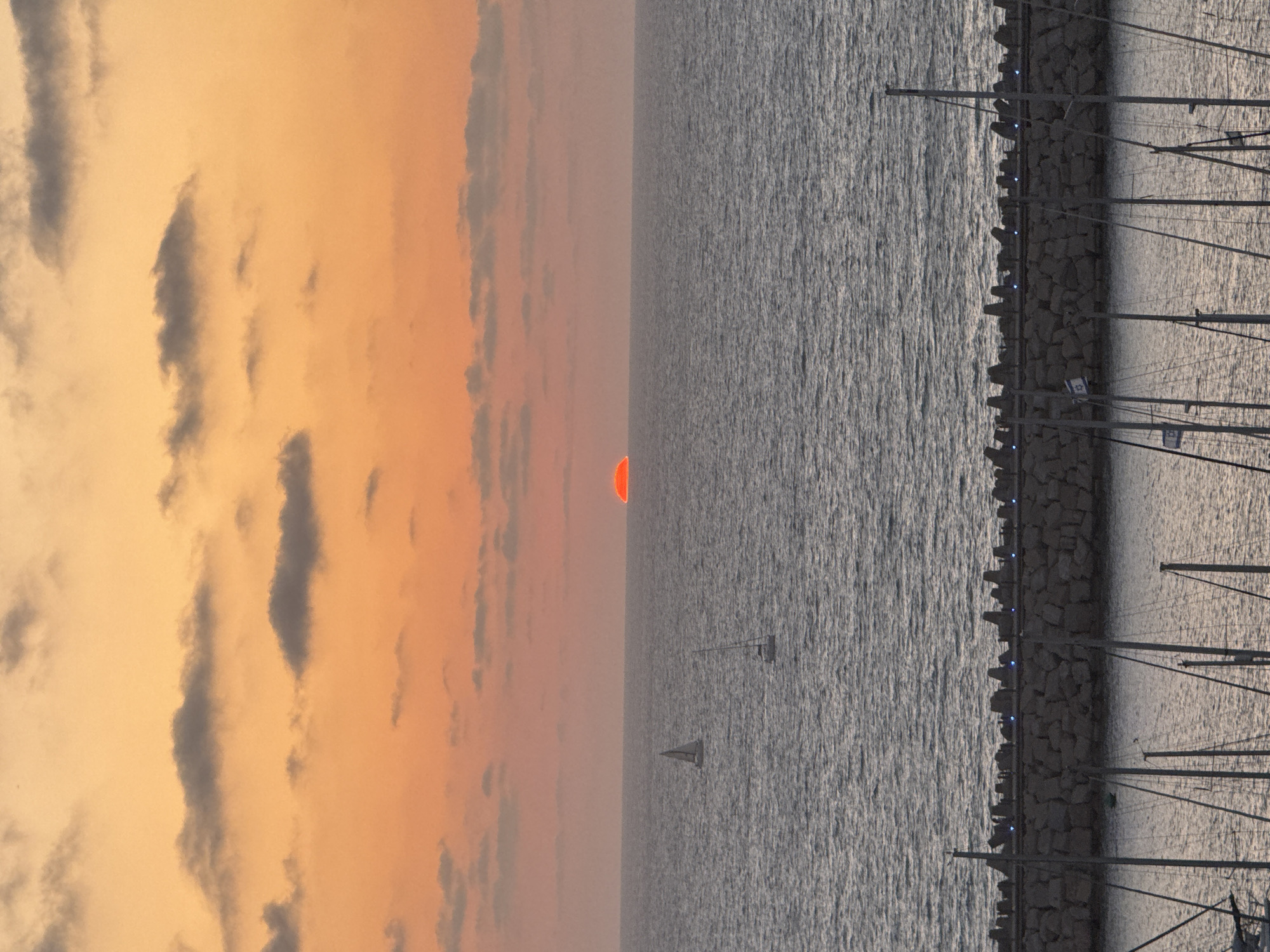 The image depicts a serene sunset or sunrise scene over the sea, likely at Herzliya Marina, Israel. The sky, dominating the left portion of the frame, is bathed in warm orange and peach tones, interspersed with scattered dark grey clouds. A perfectly circular, vibrant orange-red sun is visible on the horizon line where the sky meets the water.
The right side of the image features the textured, rippled surface of the sea, appearing in shades of dark grey and silver. Two small sailboats with white sails are seen on the open water in the distance, their hulls and masts silhouetted. A dark, rocky breakwater or marina wall extends horizontally across the middle of the frame, separating the open sea from the marina area. Small, glowing blue lights are spaced along the top of this wall.
To the right of the wall, within the marina, numerous masts and rigging of moored sailboats create intricate dark silhouettes. Among these, two Israeli flags, clearly displaying the blue Star of David, are visible on separate boat masts, providing a specific location cue. The image evokes a peaceful and tranquil atmosphere at either dawn or dusk, with the artificial lights on the breakwater suggesting the latter.