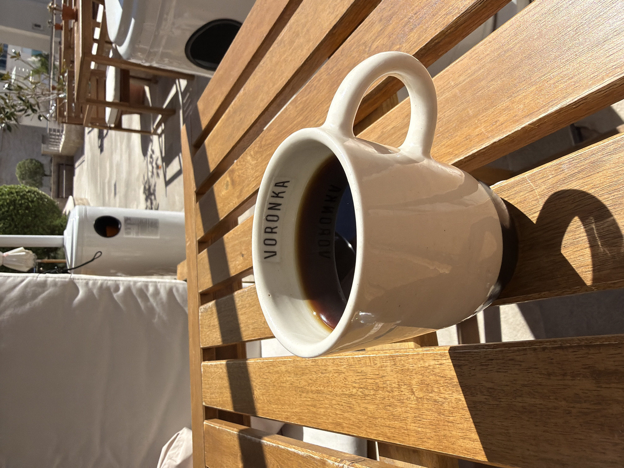 An outdoor scene features a white ceramic mug, filled with a dark liquid (likely coffee or tea), resting on a slatted wooden surface. The mug is positioned at an angle, with its handle visible to the right. Inside the mug, along the rim, the word "VORONKA" is clearly visible, with its reflection also seen in the liquid.
The setting is bathed in bright sunlight, creating strong, defined shadows from the mug and the wooden slats, indicative of a clear day, possibly morning or late afternoon. The wooden surface, which appears to be part of an outdoor table or furniture, shows some signs of wear.
In the background, there's a glimpse of an urban or semi-urban environment with buildings under a bright sky. Greenery, resembling manicured bushes or small trees, adds natural elements. A white outdoor appliance, possibly a heater, is partially visible to the left, alongside a white cushioned outdoor furniture piece. The scene evokes a peaceful, relaxed moment in Boreti, Montenegro. No specific activity is depicted beyond the mug resting on the table.