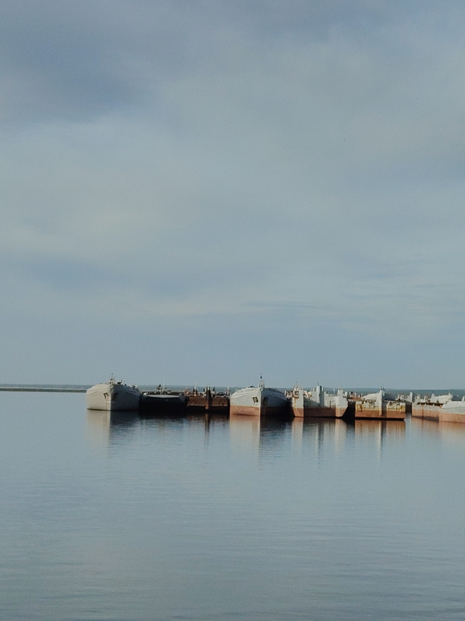 The image depicts several ships or barges docked or moored in calm water, possibly a harbor or bay, under a cloudy sky. The vessels vary in color and design. In the background, a thin strip of land can be seen, but there is no information to identify the location.