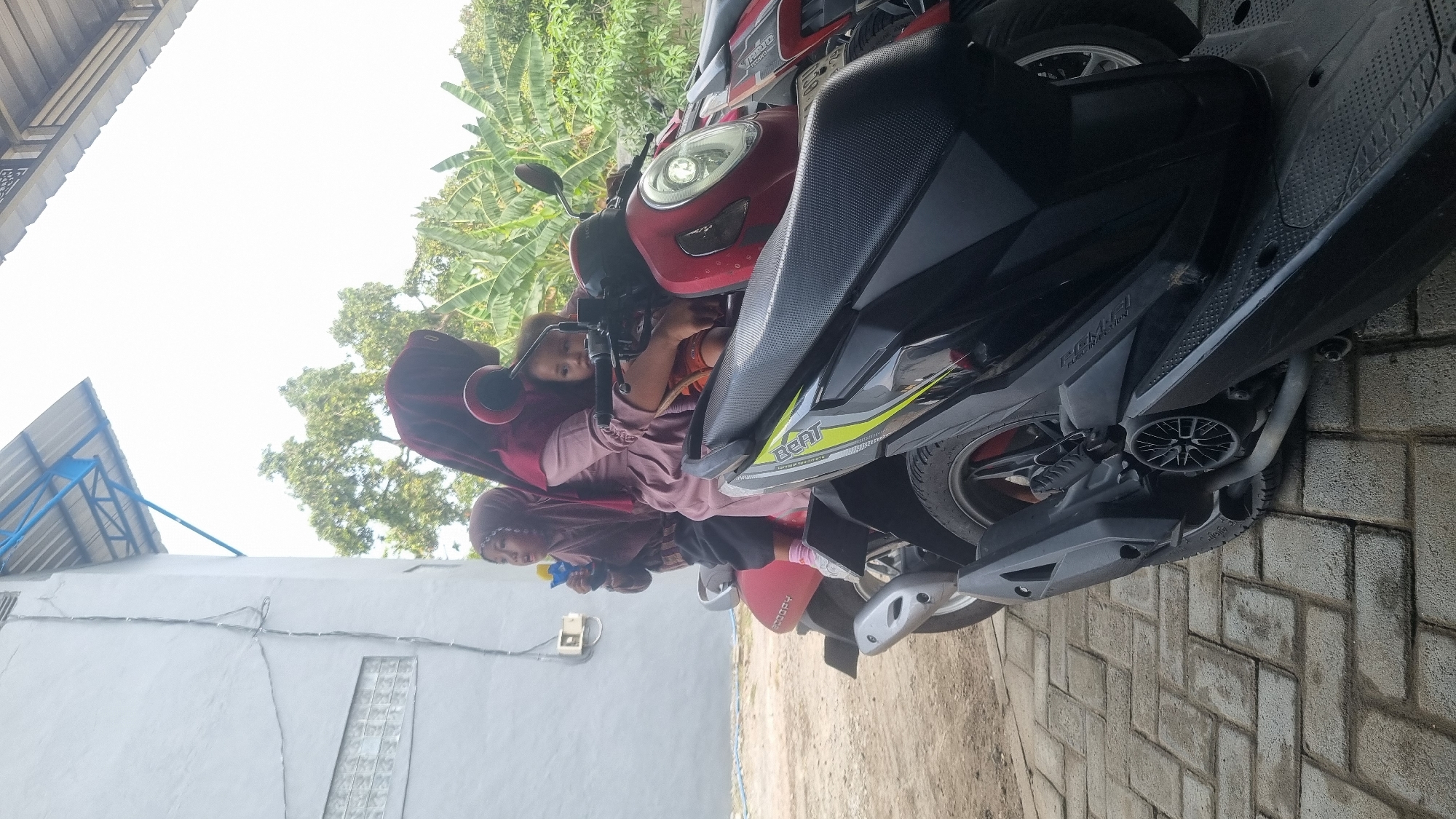 In Diwek, Indonesia, a woman wearing a maroon hijab and a pink dress sits on a red scooter, holding two young children. One child, a girl, is seated in front of her, wearing a brown hijab and a white top with pink leggings, and is holding a blue toy. Another child, a boy, sits behind the woman, wearing an orange shirt. The scooter is parked next to a black scooter, and they are both on a paved area with a brick pattern. Behind them, a white building with a blue roof structure is visible, along with lush green trees and foliage. The scene appears to be outdoors, possibly in a residential or public area.