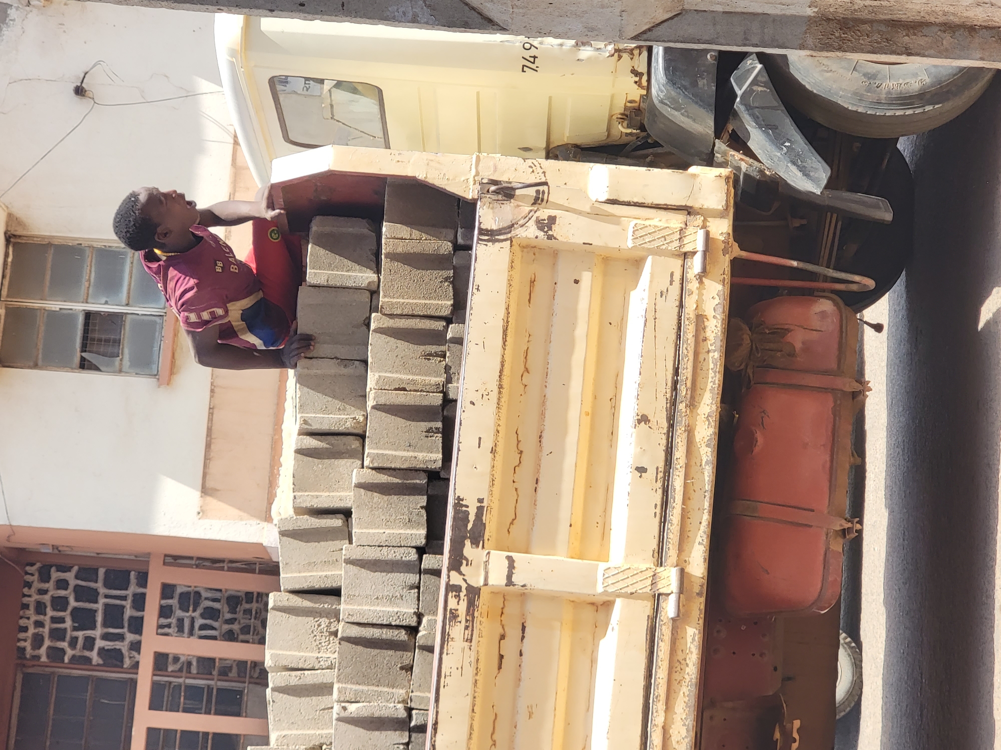 A young boy is partially visible, perched on a stack of gray rectangular blocks, reaching towards the cab of a light yellow truck. The truck is parked beside a building in Maiburiji, Nigeria. The building has windows with grills. The truck's bed appears to be metal and shows signs of rust. A red tank is affixed to the side of the truck.