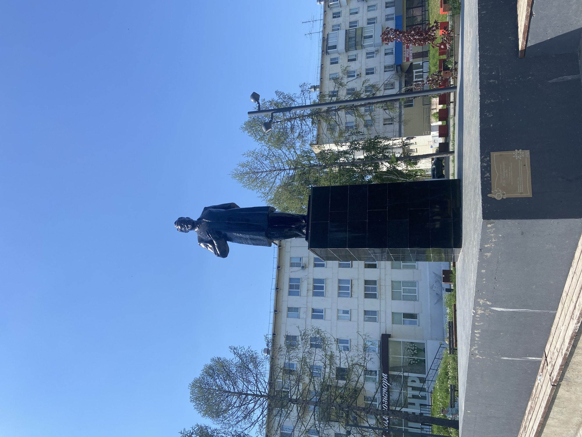 The image shows a statue on a black, tiled pedestal. The statue is dark and appears to be of a male figure in a long coat. Behind the statue is a multi-story building with numerous windows. There is also a streetlamp and some trees in the background. To the right of the pedestal is a plaque affixed to a darker grey surface. The environment appears to be an urban setting with clear, blue skies.
The text on the storefront behind the statue suggests that the location is the city of Chaikovsky, Russia.