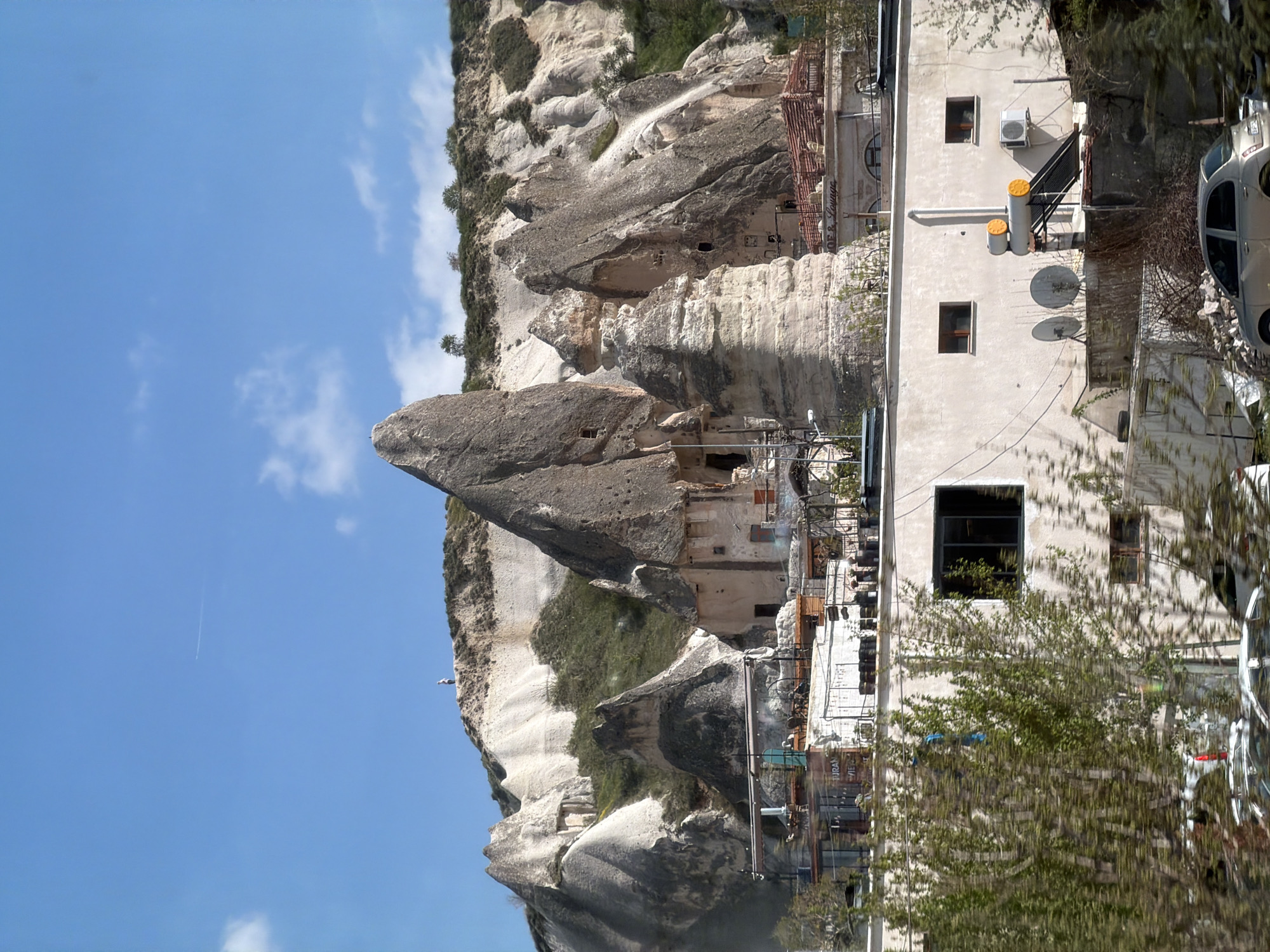 This image captures a vibrant daytime scene in Göreme, Cappadocia, Turkey, showcasing its distinctive landscape of "fairy chimney" rock formations interspersed with human settlements. The sky is a clear blue with scattered white clouds, and a faint airplane contrail is visible in the upper left.
The primary subjects are the towering, conical rock formations, or fairy chimneys, which dominate the left and central parts of the frame. These natural structures exhibit textures indicative of erosion and also reveal numerous carved-out cave dwellings, with visible windows and entrances. Patches of green vegetation cling to the sides of these rocky hills.
To the right, a modern-style, light-colored building stands in contrast to the natural rock. This structure features several rectangular windows, an external air conditioning unit, two yellow cylindrical objects (likely solar water heaters), and multiple satellite dishes. A staircase with a dark railing provides access to an upper level. A light-colored car is partially visible, parked on the far right.
Further into the scene, additional buildings and structures are integrated into the rock formations, some with terracotta-colored roofs. Visible text on a sign or awning reads "Lounge," suggesting a hospitality establishment. Below this, another partial text appears to be "URAN." The image portrays a harmonious blend of unique natural geology and human adaptation, under bright, sunny conditions.